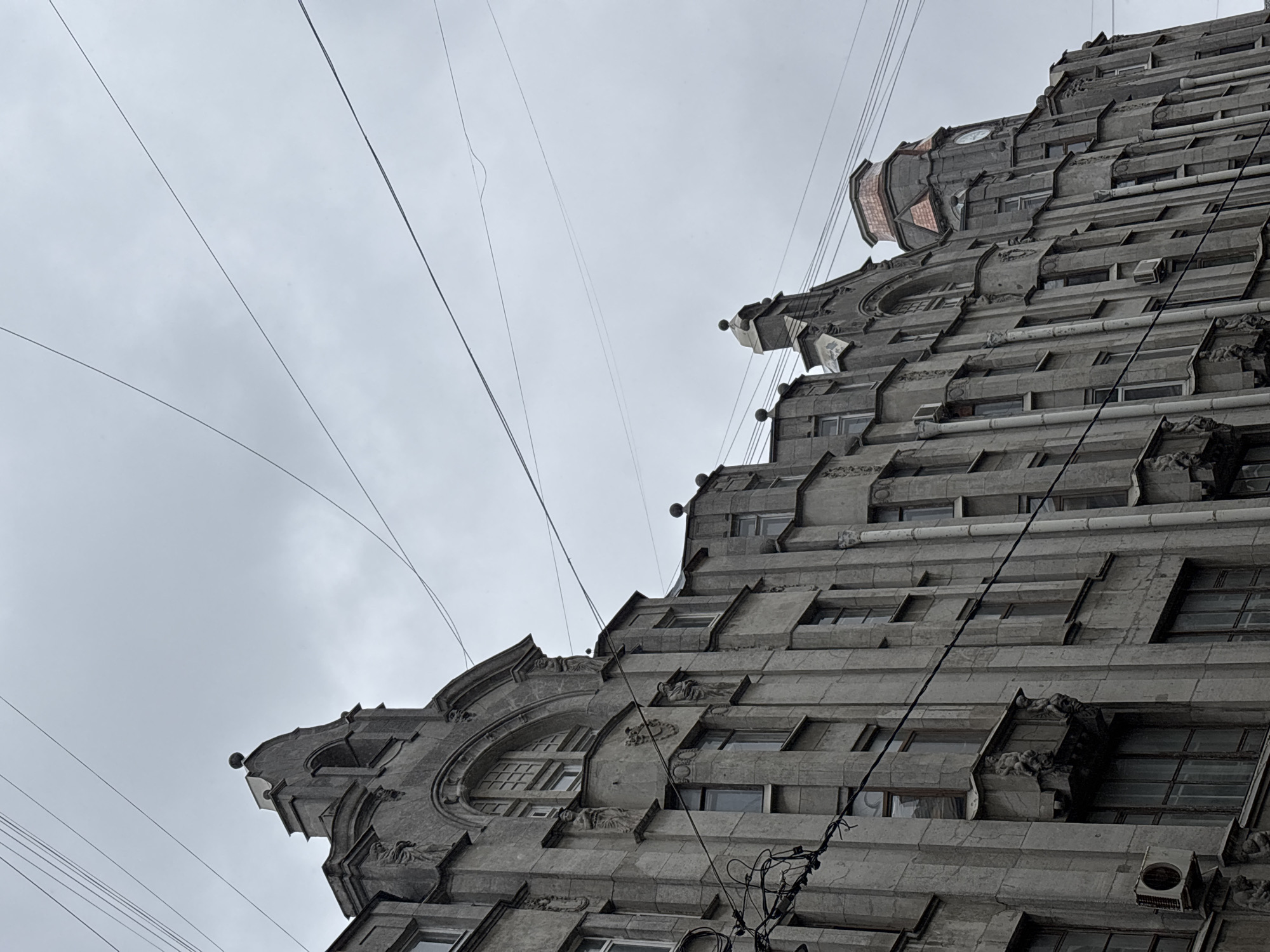 A low-angle photograph captures a multi-story building in Saint Petersburg, Russia, distinguished by its gray stone facade and eclectic architectural details. Multiple windows are visible, integrated into the building's geometric design, and intricate carvings adorn its surface. The cloudy sky serves as a backdrop, pierced by several overhead power lines. An air conditioning unit is mounted on an exterior wall of the building.