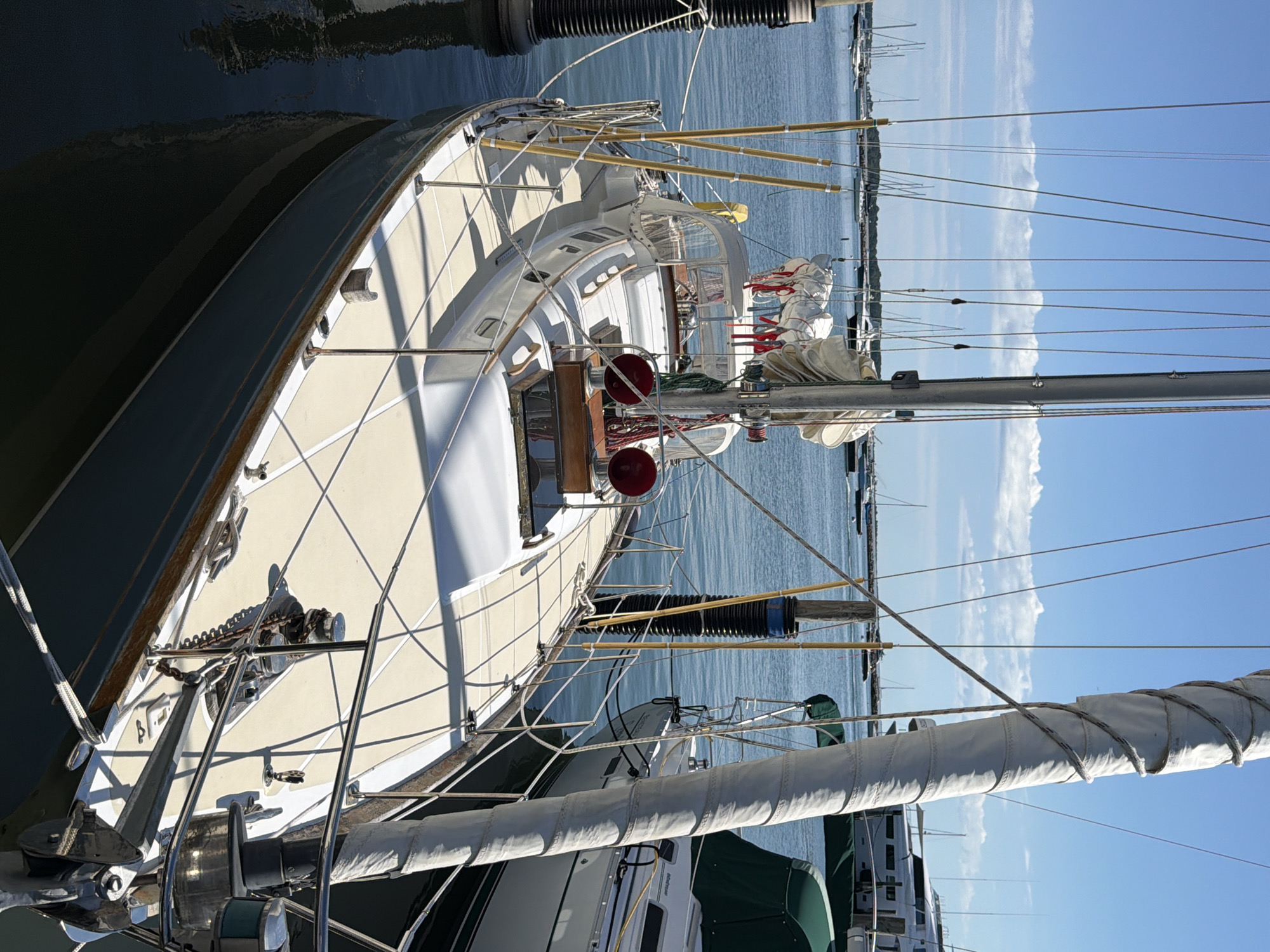 The image shows a sailboat docked in the Village of Sag Harbor, United States of America. The sailboat is the primary subject, featuring a white hull with a blue stripe along the waterline, a beige deck, and a tall mast. Several lines and sails are visible, some furled and tied. The boat is moored next to a dock, with pilings and ropes securing it. In the background, other boats and a large white yacht are visible on the water under a clear blue sky with scattered clouds. The overall scene depicts a peaceful harbor setting on a sunny day. There are no people visible in the image.