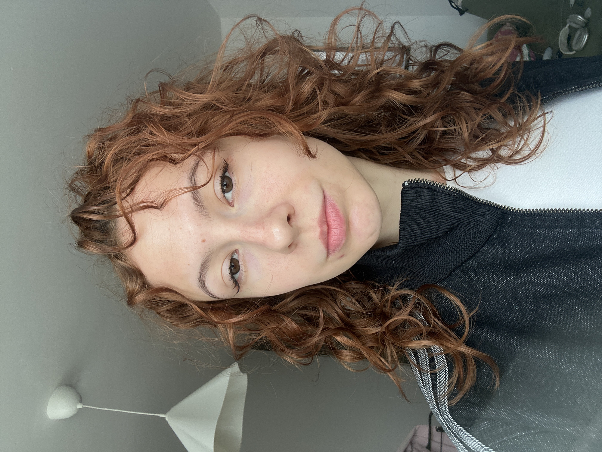The image displays a young person with fair skin and abundant, long, reddish-brown curly hair, lying down and looking directly into the camera. Their brown eyes are open, and their lips are closed with a natural, subtle pink hue and a slight sheen. Faint freckles are visible across their nose and cheeks.
The person is wearing a dark, possibly black or navy blue, athletic jacket with a visible zipper down the front. A white garment is worn underneath. The jacket features distinct grey and white stripes on one side, characteristic of athletic wear. The voluminous curly hair frames the face and spreads out around the head, occupying a significant portion of the frame.
The setting is indoors, appearing to be a casual room during daytime, suggested by the natural lighting. Above the person's head, a light grey ceiling is visible, from which a white, bell-shaped lampshade or light fixture hangs by a white cord. In the upper background, some blurred, out-of-focus shapes are discernible, possibly including headphones, indicating other objects in the room. The person's expression is calm and neutral, suggesting a relaxed, posed moment. There is no visible text in the image. While the provided context states the location as Novi Sad, Serbia, there are no specific visual cues within the image itself to confirm this geographical location.