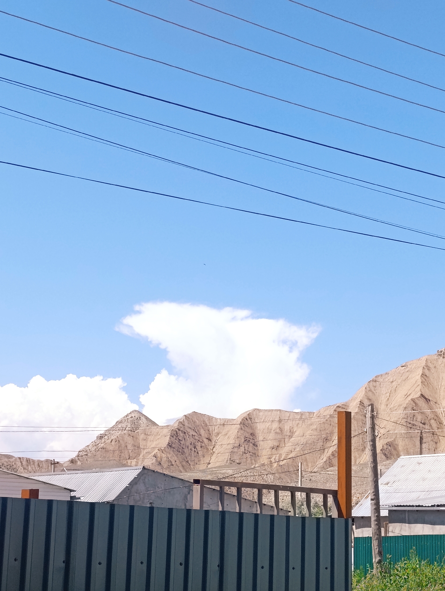 In Naryn City, Kyrgyzstan, an outdoor scene unfolds under a clear blue sky with scattered white clouds. Power lines stretch horizontally across the upper portion of the frame. Below them, rugged, tan-colored mountains dominate the background, their surfaces bearing the marks of erosion. In the mid-ground, various structures suggest a developing or inhabited area. A dark green corrugated metal fence runs along the bottom, partially obscuring the view of buildings behind it. These buildings feature corrugated metal roofs and a mix of lighter-colored walls. A rusty metal pole stands vertically near the center right, next to a wooden utility pole. The overall impression is of a semi-rural or developing urban fringe in a mountainous region.