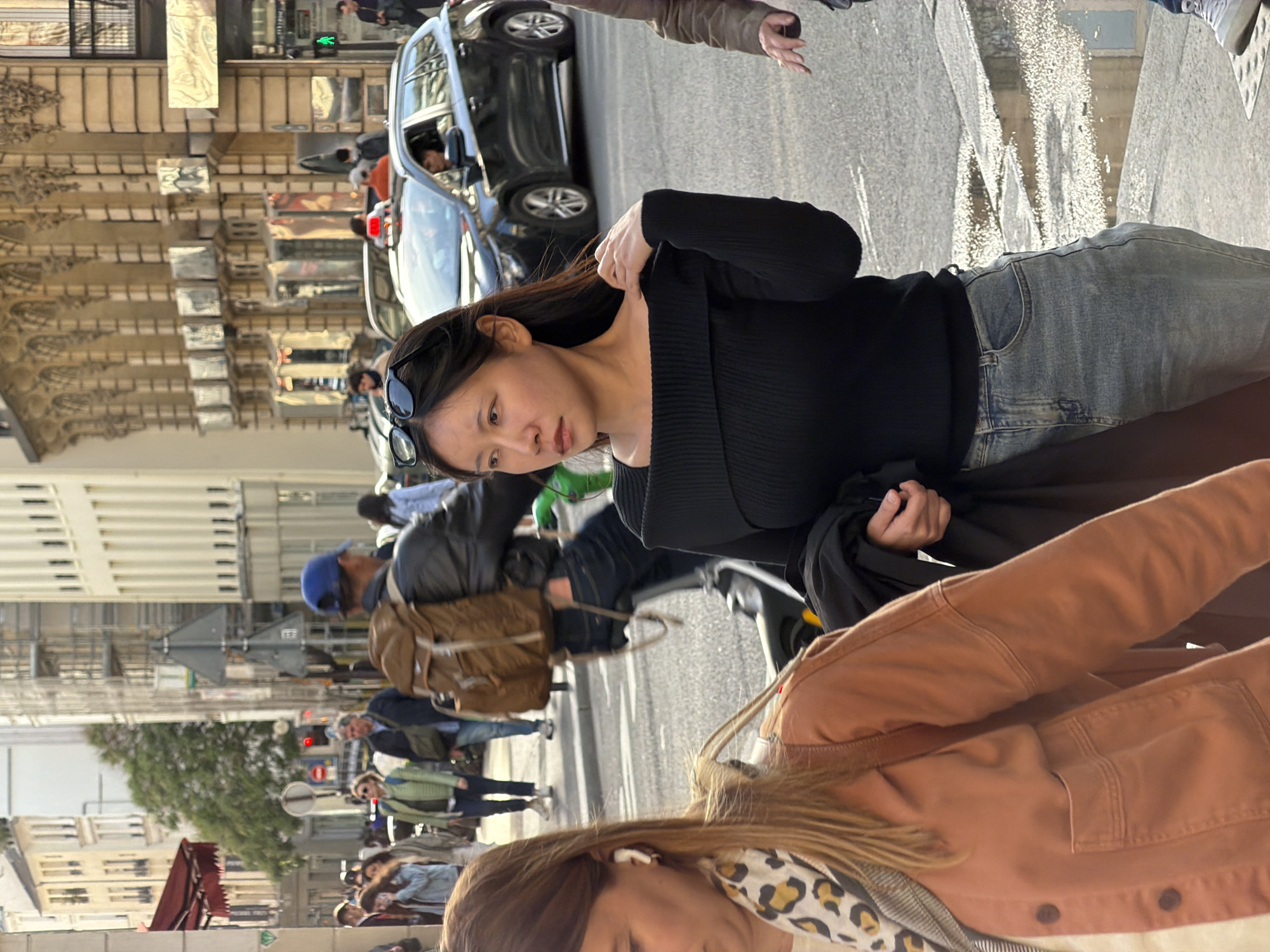 This medium close-up, rotated 90 degrees clockwise, captures a daytime street scene in Paris, France. The primary subject is an East Asian woman positioned in the center, facing slightly left. She has long dark hair, wearing a form-fitting black long-sleeved top and light blue denim jeans, with black sunglasses resting on her head. Her expression is neutral to contemplative as she looks off-frame.
Partially visible in the lower right corner is another person with light brown or blonde hair, wearing a rust-colored jacket and a black and tan leopard print scarf, with white earbuds in their ears.
The background reveals a bustling city street. A dark-colored SUV is prominent in the mid-ground, driving on the paved road. Further back, various pedestrians and other vehicles are visible. Classic Parisian architecture lines the street, featuring multi-story buildings with ornate facades and intricate detailing. A green pedestrian traffic light can be spotted in the distance amongst the buildings, confirming urban activity. Other individuals in the background include a person wearing a blue cap and a brown backpack, seemingly on a bicycle.
The lighting suggests a bright, clear day, likely late morning or early afternoon. The overall atmosphere is that of a lively urban environment. No discernible text is visible.