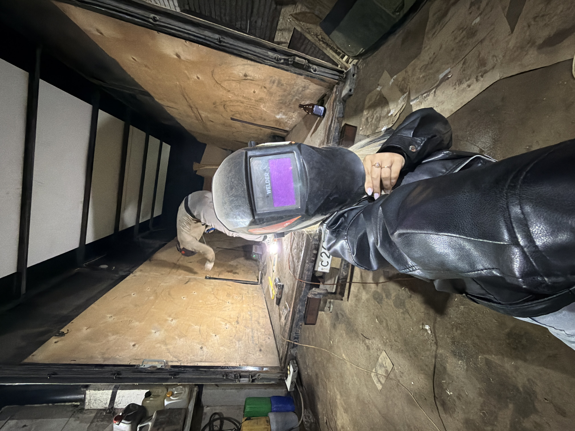 The image depicts an indoor industrial or workshop setting, likely in Chaikovsky, Russia, captured from a first-person perspective at an oblique angle. Two individuals are visible. In the foreground, a person is wearing a black welding helmet, which has the word "WELDER" displayed on its purple auto-darkening screen. This individual also wears a black leather jacket, and their hand, adorned with rings and light-colored nail polish, is visible near their chest, suggesting they are holding the camera.
In the mid-ground, a second person is actively engaged in welding, indicated by the bright light and sparks emanating from their work. This individual wears light-colored work clothes, possibly a jumpsuit or overalls, and is positioned further down the length of the working area.
The environment features a mix of materials: a light brown plywood platform or floor on the left, and a rougher, darker concrete or dirt-like floor on the right. Various objects are scattered throughout the scene, including a brown glass bottle on the wooden surface, several wires and cables, and some tools. A metal plate or object marked "C2" is visible near the foreground figure. In the lower left corner, a group of colorful containers (green, blue, and yellow) are stacked. On the far left, a series of white vertical panels with dark supports line the wall. The lighting is primarily artificial, with the welding arc providing a significant light source.