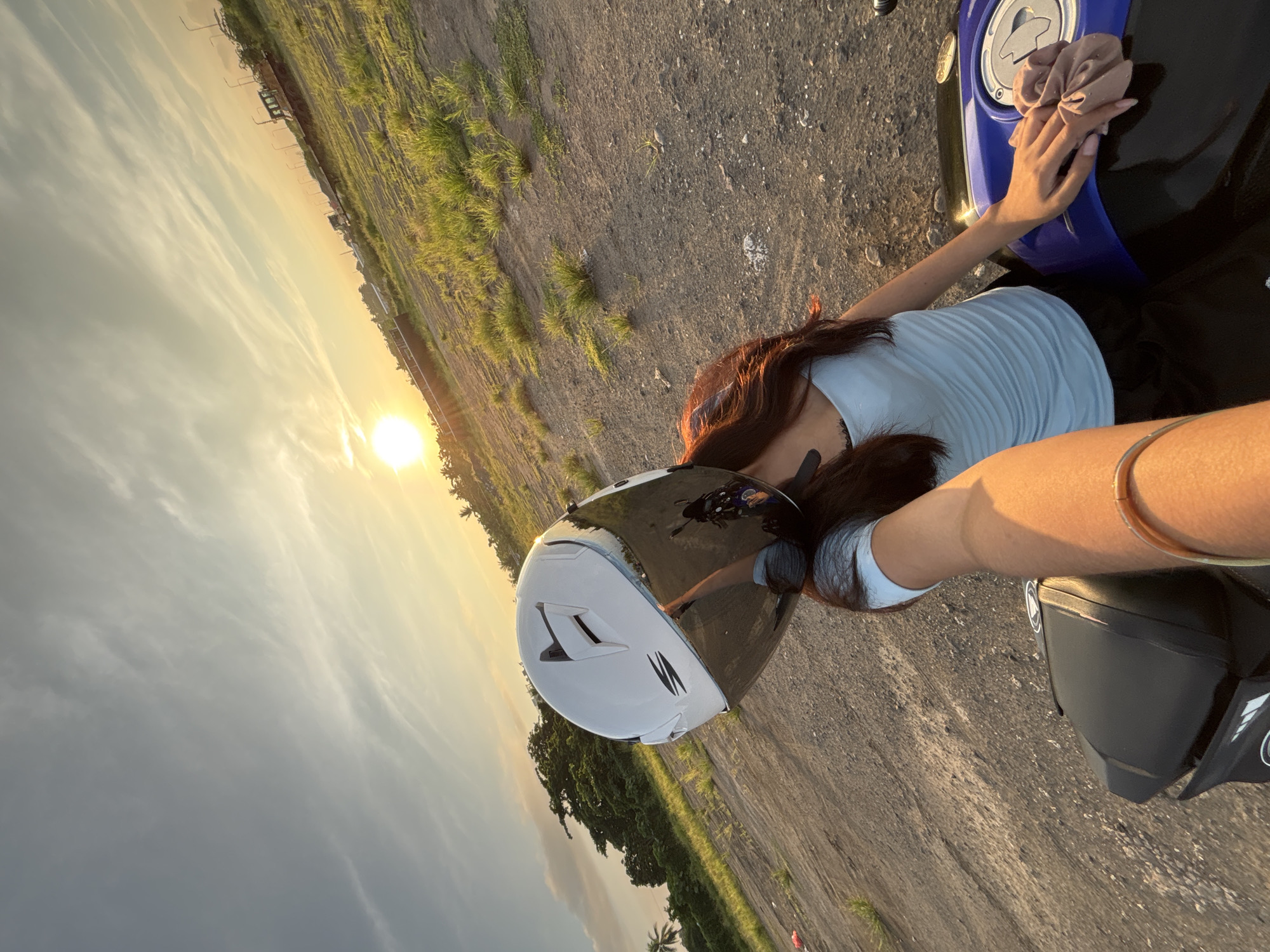 A person wearing a white motorcycle helmet with a reflective visor is seated on a black and blue motorcycle. The person has long, dark hair and is wearing a light blue tank top and black pants. The helmet's visor reflects the image of the person and the motorcycle. The scene appears to be an open, dusty area with dry grass and some trees in the background. The sun is low in the sky, casting a warm glow and long shadows, indicating it is either sunrise or sunset. In the distance, there are some structures and what appear to be streetlights or power lines. This setting is consistent with a rural or undeveloped area on the outskirts of Davao City, Philippines, possibly a vacant lot or a construction site. The overall atmosphere is casual and relaxed, with the person appearing to be taking a selfie or being photographed.