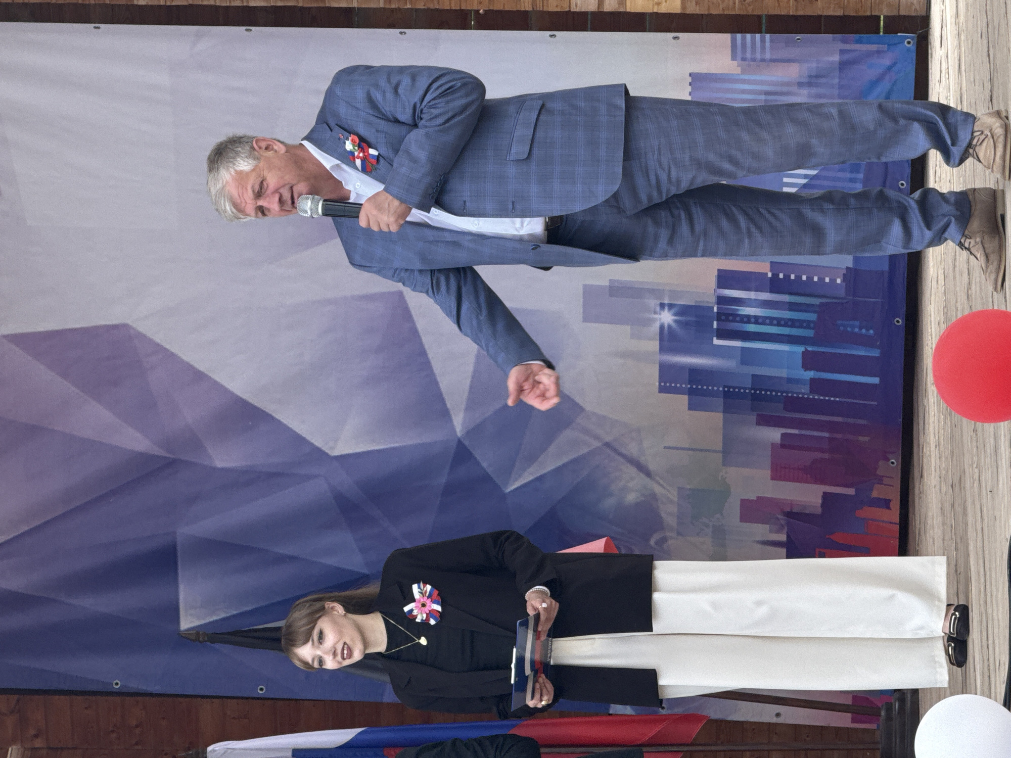 In the city of Votkinsk, Russia, a woman and a man stand on a wooden stage in front of a blue and purple banner depicting abstract geometric shapes and a city skyline. The woman, on the left, wears a black blazer over a black top and white wide-leg pants. She holds a clipboard and a document with red and white stripes. A ribbon decoration with pink, white, and blue elements is pinned to her blazer. She has blonde hair with bangs and is looking to her left. The man, on the right, is in a blue plaid suit with a white shirt. He holds a microphone in his right hand and appears to be speaking, with his left arm bent and his hand open. A red, white, and blue ribbon decoration is pinned to his suit jacket. The man has short, graying hair. In the foreground, two large balloons are visible: a white one on the left and a red one on the right. Russian flags are positioned behind the woman on the left.