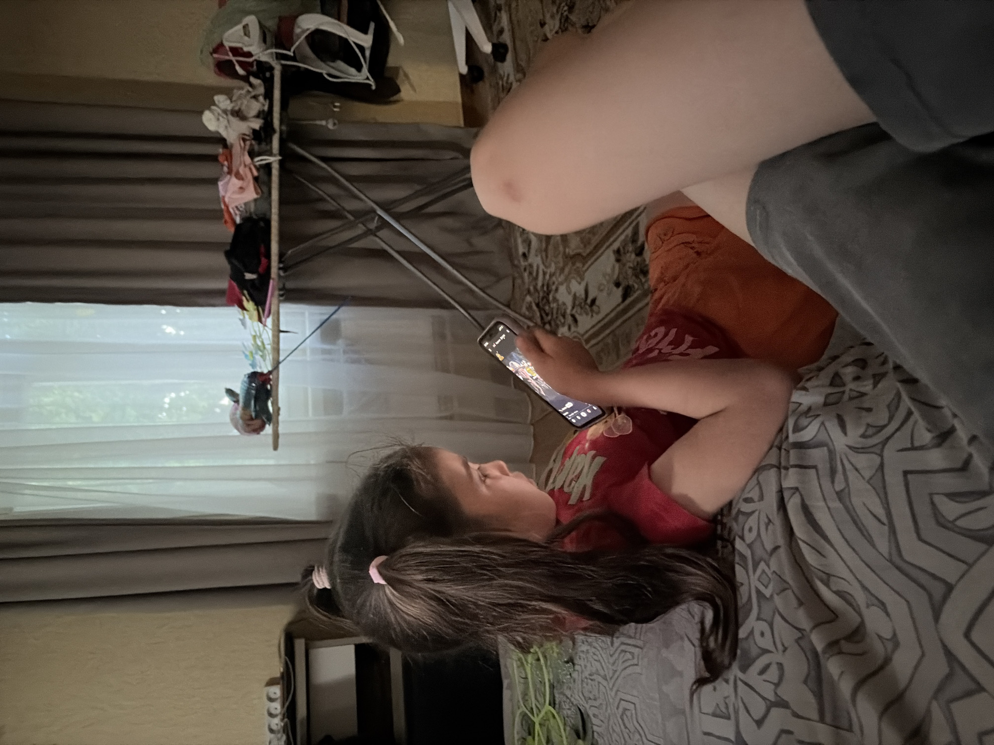 A young girl with dark hair tied in ponytails is lying on a bed, holding a smartphone and looking at its screen. She is wearing a red t-shirt. Another person, likely an adult, is partially visible in the foreground, their legs are resting on the bed. The room appears to be a bedroom, with a patterned comforter on the bed and curtains in the background. A clothes rack with various items of clothing is visible near the window, suggesting laundry may have been hung to dry. The lighting suggests it might be daytime, but the blinds are drawn, making it appear dim. The girl's focus on the phone indicates she is engaged in an activity like playing a game or watching a video. No discernible emotions are evident beyond her concentration on the device. The location context is Tambov, Russia. There is no visible text in the image.