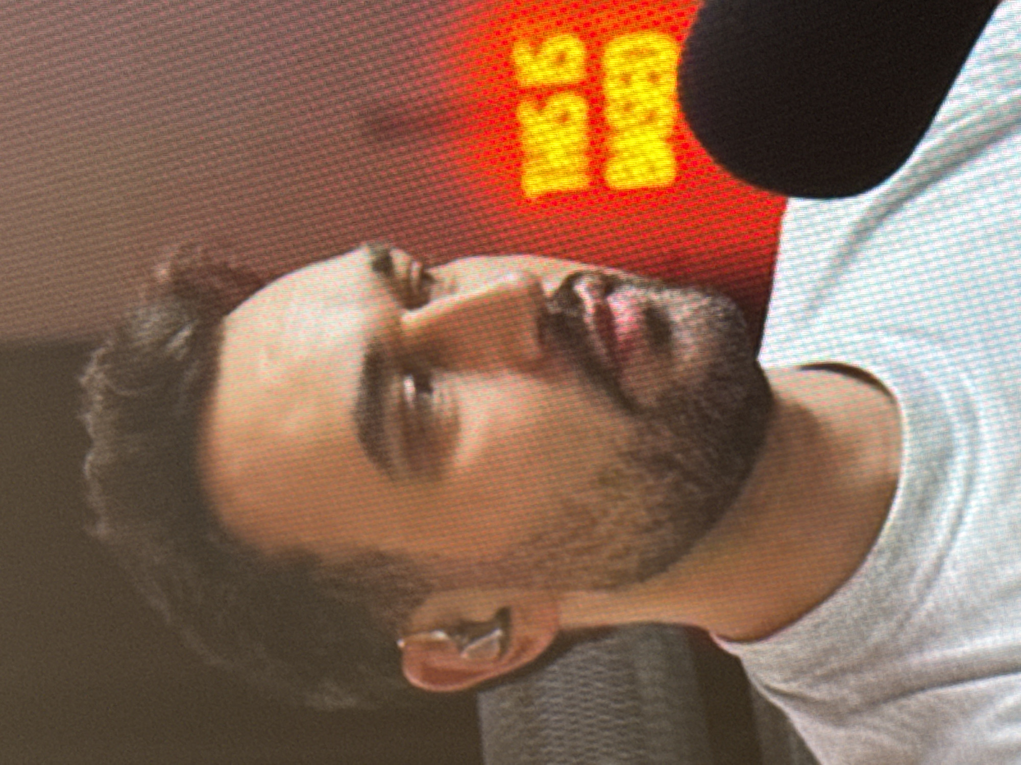 This media file features a close-up, side-profile shot of a person's head and upper body, oriented vertically in the frame. The person appears to be male with short, dark hair and stubble on his face. He is wearing a light-colored t-shirt, and in his left ear, he has a small, metallic earbud or hearing aid.
The background is blurred but shows a bright red surface with yellow text. The text is visible in the upper left quadrant of the image and appears to read "THIS IS BASED" in block letters. To the right of the text, a dark, rounded object, possibly a microphone, is partially visible. The red background suggests an indoor setting, perhaps a studio or a room with a lit display. The lighting is focused on the subject, with the red background providing a strong contrast. The image quality is somewhat pixelated, indicating it might be a screenshot or a low-resolution photograph. There are no immediate cues to suggest the time of day or weather conditions, and the emotion of the subject is not clearly discernible due to the angle and focus. The location context of Saratov, Russia, is not visually indicated within the image itself.