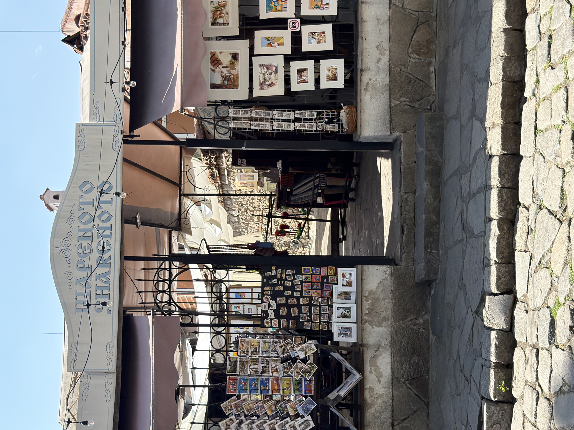This image captures the outdoor entrance of a shop named "ШАРЕНОТО ШАРНОТО" (Sharenato Sharnoto), located in the Old Town of Plovdiv, Bulgaria, under bright daylight conditions.
The shop's facade features a prominent, light-colored sign with stylized blue Cyrillic text and decorative scrollwork. A string of incandescent bulbs is suspended horizontally across the sign. Below the sign, two extended awnings, one tan (left) and one darker brown/grey (right), provide shade. The entrance itself is an open, dark metal gateway, partially adorned with decorative ironwork, leading into a deeper, less distinct interior space with visible stone walls and additional awnings.
To the left of the entrance, a multi-tiered display rack holds numerous postcards featuring various images. Adjacent to this, a dark vertical panel exhibits a collection of smaller framed pictures or magnets. Below these, four larger, horizontally oriented framed images rest on a stone ledge.
To the right of the entrance, a vertical stand displays multiple framed artworks, predominantly paintings or watercolors depicting architectural and street scenes. A circular red and white "no photography" symbol is partially visible among these frames. A small, woven basket containing a white, fluffy object rests on the lower ledge.
The ground in front of the shop is a textured surface composed of irregular flagstones and rectangular cobblestones, with minimal grass patches between them. A clear boundary of rectangular stones separates the immediate foreground from the area directly in front of the shop's elevated stone base. Several stone steps lead up from the cobblestone street to the shop entrance. The lower portion of the building's facade consists of rough-hewn stone blocks topped by a section of light, weathered plaster. No individuals are visibly present within the immediate scene.