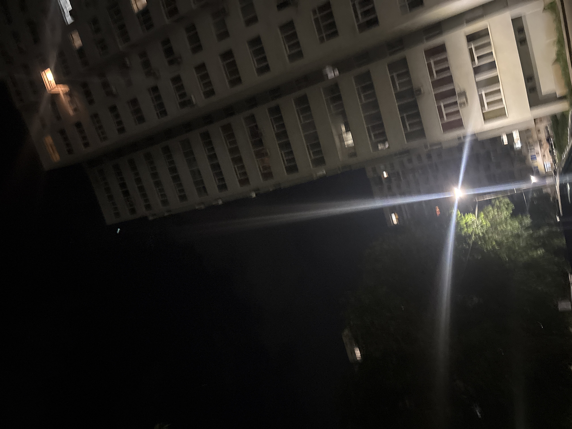 The image captures a nighttime scene of a residential building in Dobrich, Bulgaria. The building, likely an apartment complex, is tall and has numerous windows, many of which are illuminated from within, suggesting occupancy. The facade of the building is light-colored, contrasting with the dark night sky. A bright, artificial light source, possibly a streetlight or a powerful lamp, is visible in the foreground, casting a strong glare across the image and illuminating a nearby tree with green foliage. The tree appears to be located on the grounds of the building or in its immediate vicinity. In the background, another, slightly shorter building is visible, also with illuminated windows. The overall impression is one of a typical urban residential area at night. The perspective suggests the photo was taken from ground level, looking upwards at the buildings.