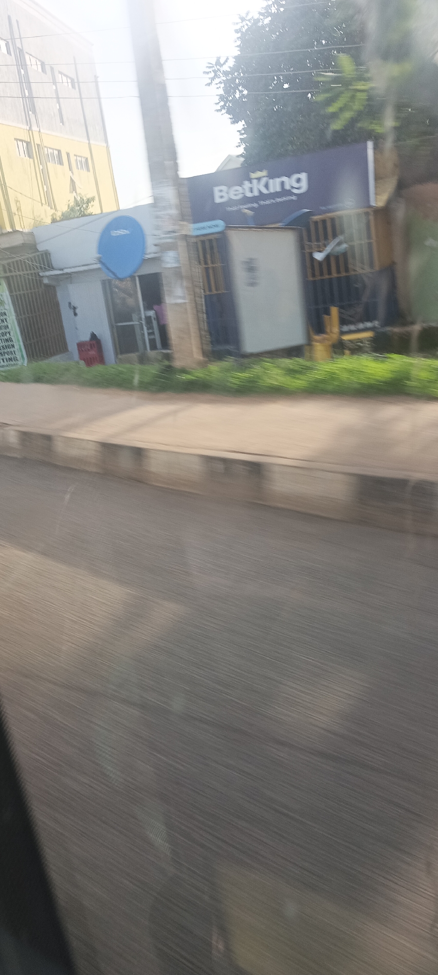 This image captures a street scene in Jos, Nigeria, during the daytime, viewed from a moving vehicle. The foreground shows a paved road with significant motion blur, indicating motion, alongside a raised concrete sidewalk and curb.
On the left side of the frame, a multi-story building with a light yellow facade and numerous windows is partially visible in the background. Closer to the road, a small, white-painted structure, likely a shop or kiosk, stands. A prominent blue satellite dish, resembling a DSTV dish, is mounted on its exterior. A red plastic crate is placed near the shop's entrance, and a faint figure can be seen inside. To the left of this shop, a green sign displays partial, blurred text, including fragments like "TION," "ANT," "COPY," and "NTING," suggesting business services.
Adjacent to this shop, on the right, is another small commercial structure with windows protected by metal grates. Outside this structure, some yellow items, possibly chairs or crates, are visible. Above these shops, a large dark blue billboard proudly displays the text "BetKing" in white, accompanied by a crown logo. Below the main "BetKing" text, fainter white text is visible, possibly reading "Welcome. Home of Kings." Trees with dense green foliage are visible behind and above the buildings against a bright, clear sky. The overall impression is a bustling roadside in an urban or semi-urban area.