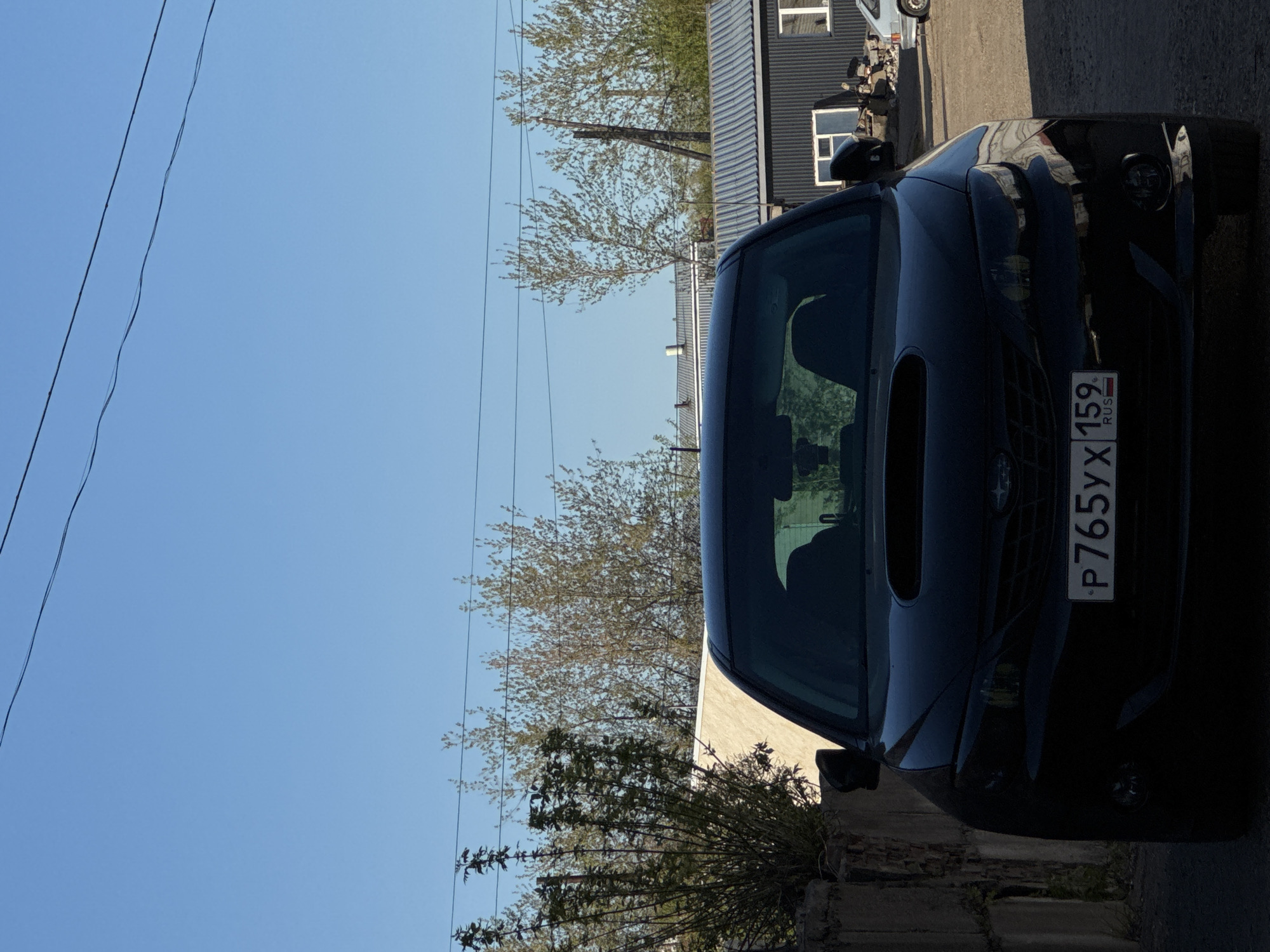 The image presents a close-up, rotated view of a dark-colored Subaru car. The car is positioned near a concrete structure, possibly a building foundation or retaining wall. A small bush or shrubbery is located near the front of the car, partially obscuring the lower portion of the vehicle. The car has a license plate that reads "P765YX 159 RUS," indicating the car is registered in the Perm Krai region of Russia.
Behind the car, there is a building with dark, possibly corrugated metal siding and a couple of visible windows. Trees with budding leaves are situated beyond the building. The sky is clear and blue.
Given the license plate information and general surroundings, the location is determined to be in Chaikovsky, Perm Krai, Russia. However, the specific address is unidentifiable. There are no people visible in the frame. The focus is on the vehicle and its immediate surroundings.
