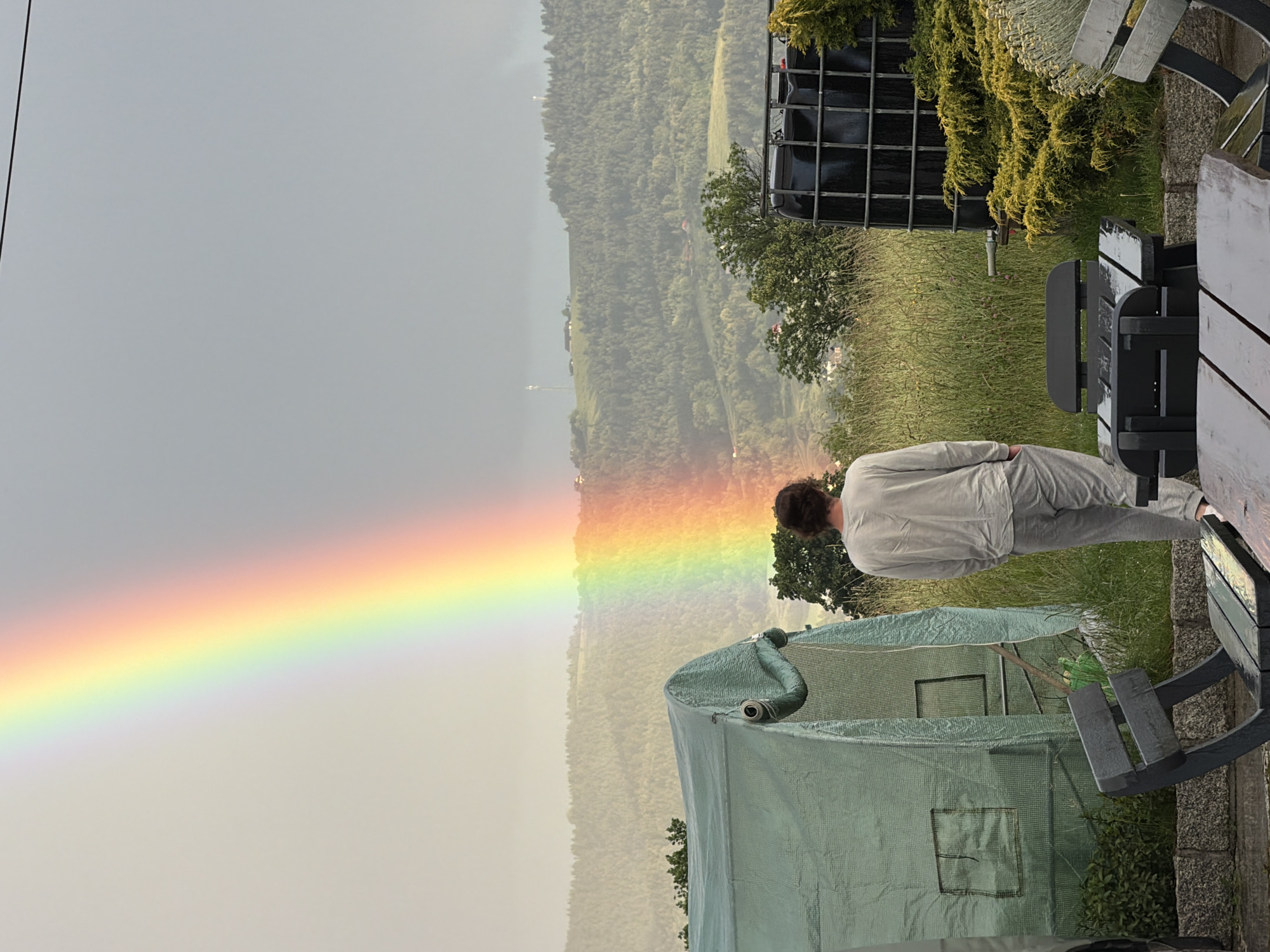 In Męcina, Poland, a person stands facing away from the camera, observing a vibrant rainbow arcing across a hazy sky. The person is dressed in light-colored loungewear, consisting of a long-sleeved shirt and sweatpants. In the foreground, a dark wooden picnic table and benches, appearing slightly wet, are visible. To the left, a green tarp-covered structure, possibly a greenhouse, is partially obscured by a large black water tank to the right. The background features rolling, tree-covered hills under a diffused, overcast sky, with scattered structures on the distant ridgeline. The overall scene suggests a rural or suburban setting following a rain shower.