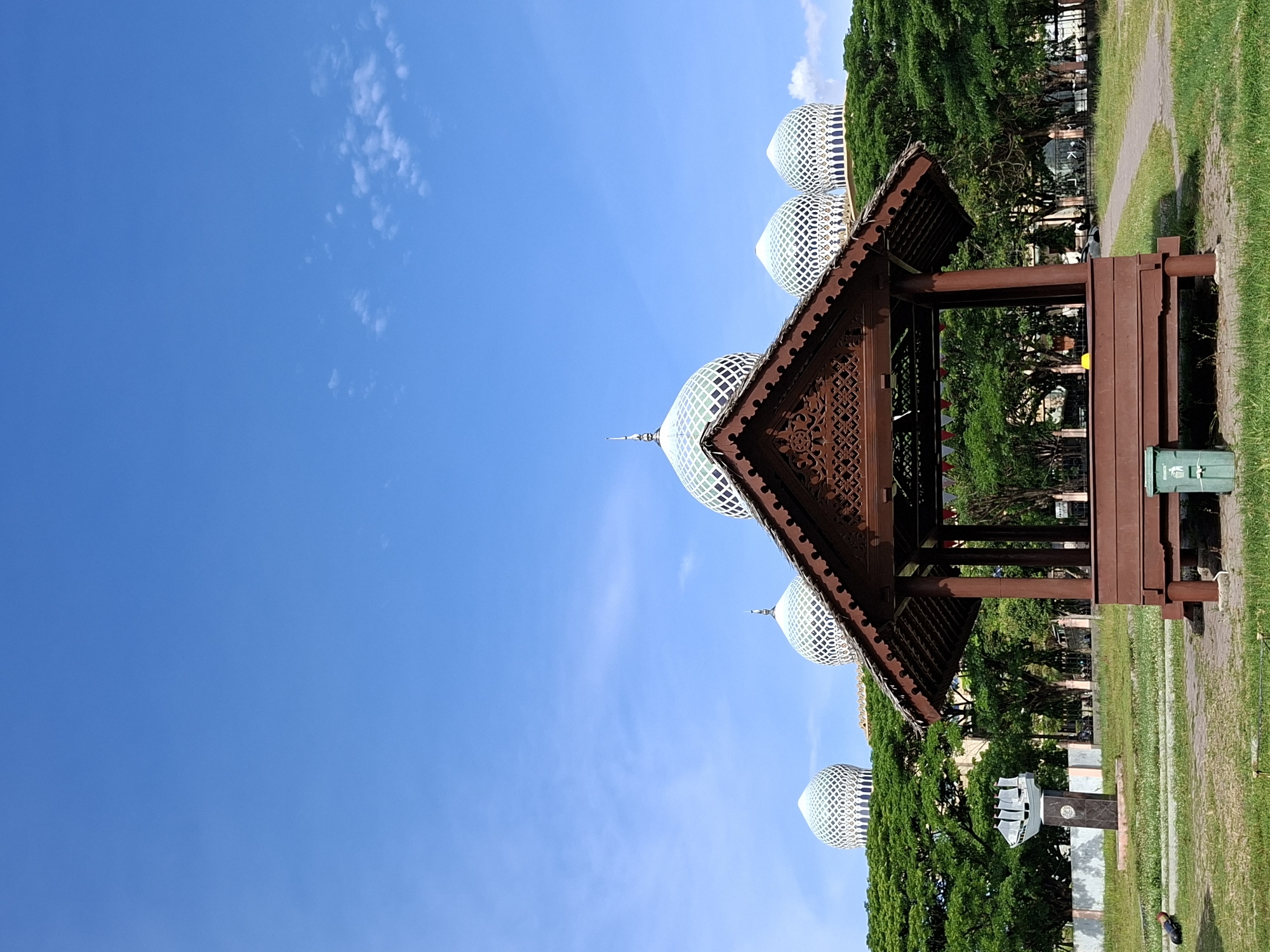 The image depicts a park setting in Lhokseumawe, Indonesia, featuring a traditional wooden gazebo with a steeply pitched roof adorned with intricate carvings. In the background, the dome-shaped roofs of the Islamic Center Lhokseumawe are visible, recognizable by their white lattice patterns. Lush green trees frame the background. The sky is clear blue with sparse clouds. The gazebo is brown, and a green trash receptacle is positioned near its side. The ground consists of green grass.