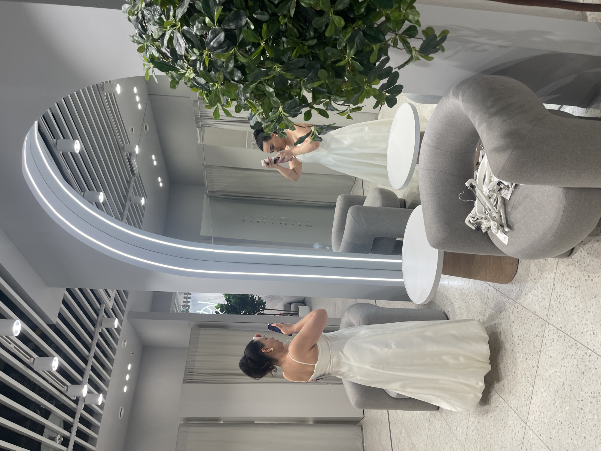 A woman, wearing a white dress, stands in a brightly lit, modern bridal shop. She is holding a phone up to her face, seemingly taking a picture or video of herself in a large, arched mirror. The shop has a minimalist design, with grey upholstered chairs, small round tables, and grey curtains. A large, leafy potted plant is positioned in the foreground on the right side of the frame. The floor is covered in light-colored terrazzo tiles. The woman has her hair pulled back in a bun and is wearing gold earrings. The mirror reflects another woman in a similar white dress, also holding a phone. The overall atmosphere is that of a sophisticated fitting or showroom. The scene is located in Moscow, Russia.