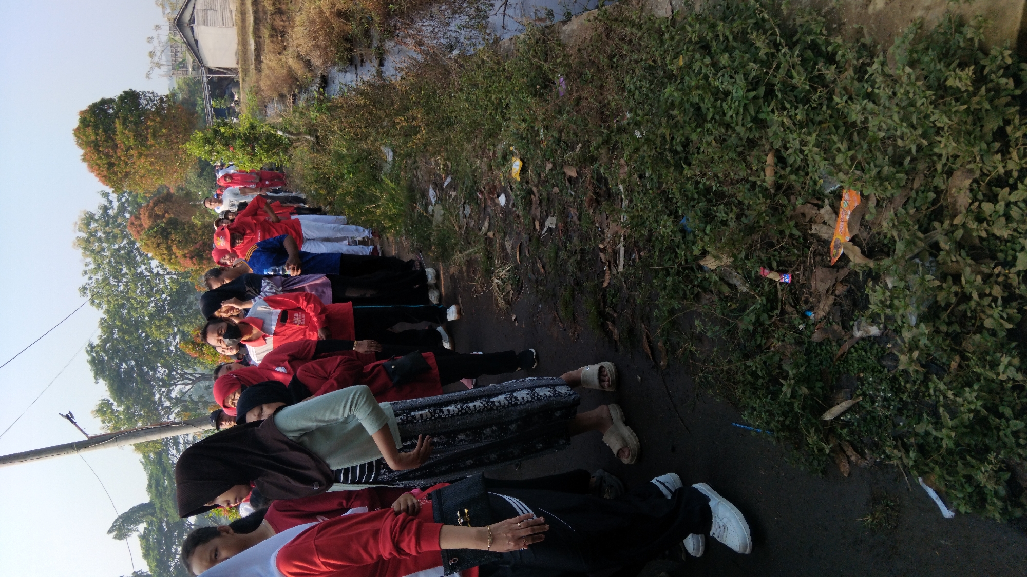 A group of people, predominantly women wearing red attire, are standing in a line along a dirt path next to a narrow canal. The path is lined with dry vegetation and scattered trash, including plastic wrappers. The people are facing to the right, with some individuals wearing headscarves. The environment appears to be a rural or semi-rural area, possibly on the outskirts of a city like Posong, Indonesia, given the presence of a small building in the background and lush green trees. The lighting suggests it is daytime, with a bright sky. The people seem to be participating in an event or activity, as indicated by their matching outfits and orderly formation.