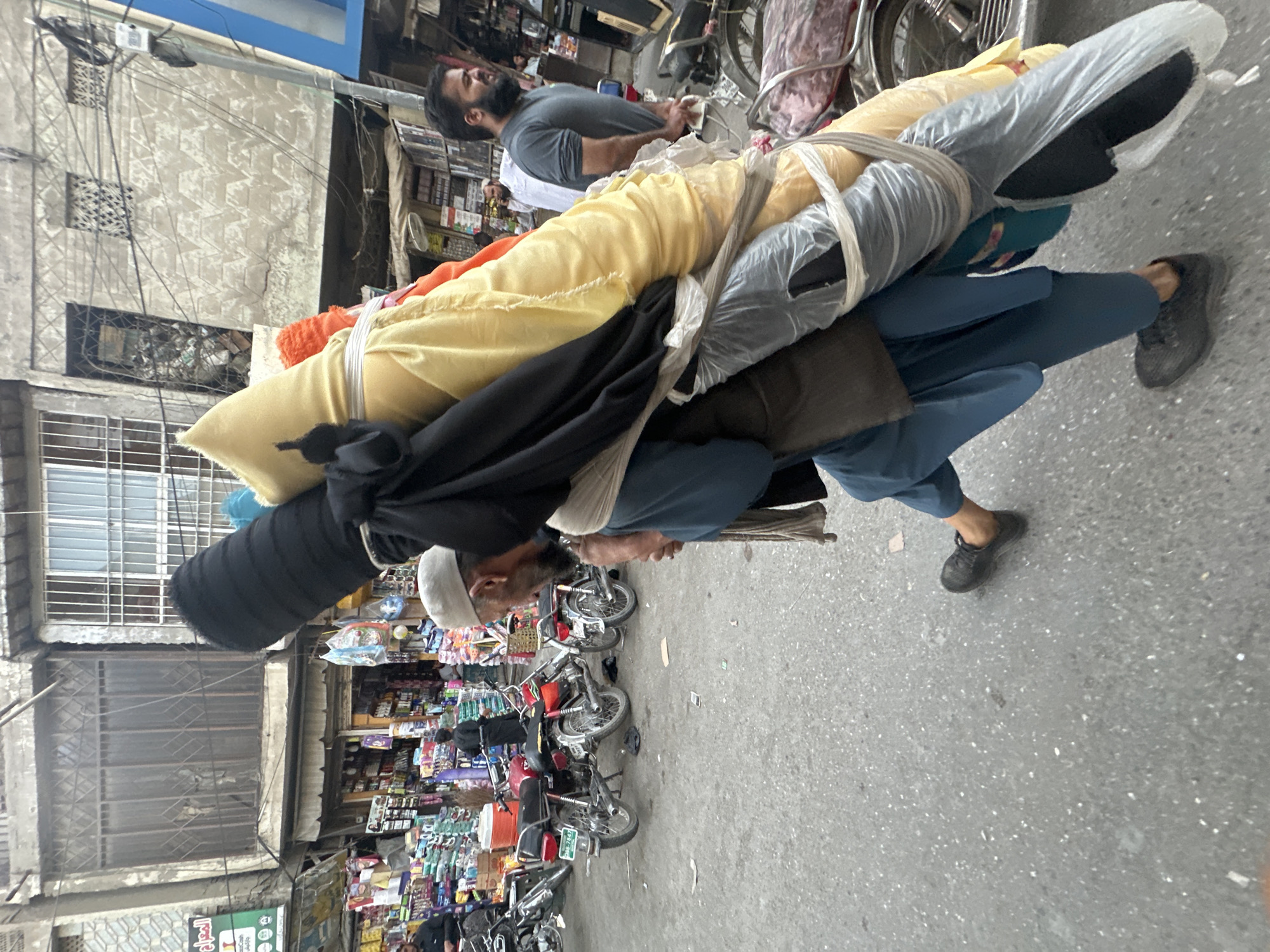 A man in Quetta City Tehsil, Pakistan carries a large bundle on his back down a street filled with motorcycles. The bundle consists of rolled fabric of various colors held together with white rope and clear plastic wrap. The man is wearing a white skullcap, black clothing, and blue pants. He is walking on a paved street next to a row of parked motorcycles. The background includes buildings with shops and residential spaces. Another man is visible behind the person carrying the load, and the scene shows signs of a busy urban environment.
