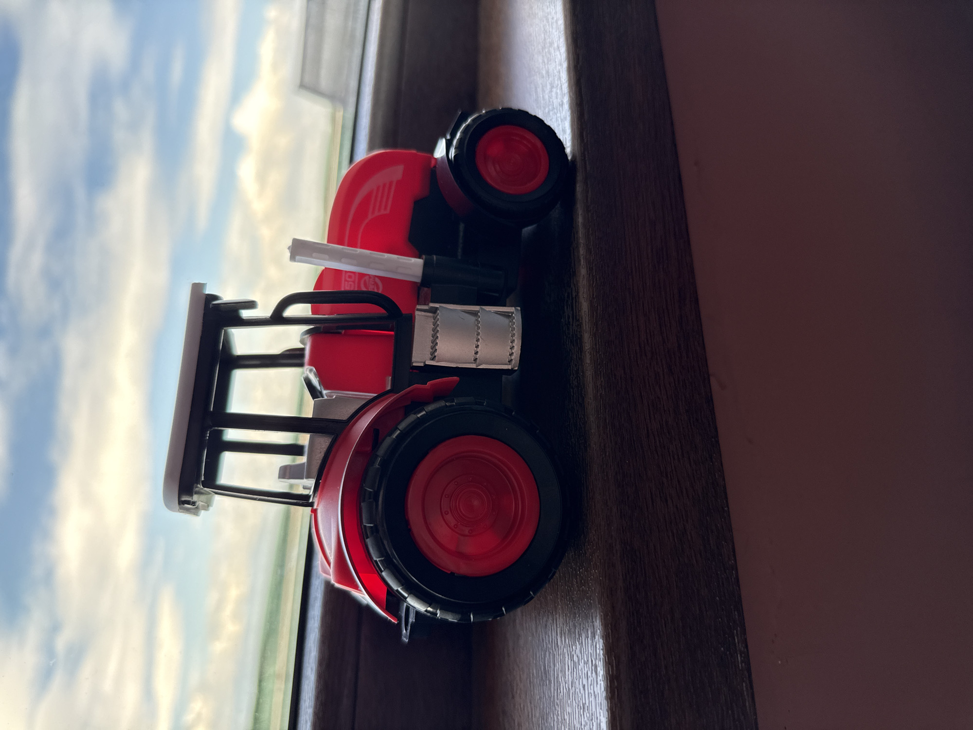 A toy tractor, red and black in color, sits on a dark brown wooden surface, likely a windowsill or shelf, with a view of the sky and possibly a field visible through a window. The sky is partially cloudy with blue patches. The location is not identifiable as Sofia, Bulgaria, based on the visual information available.
