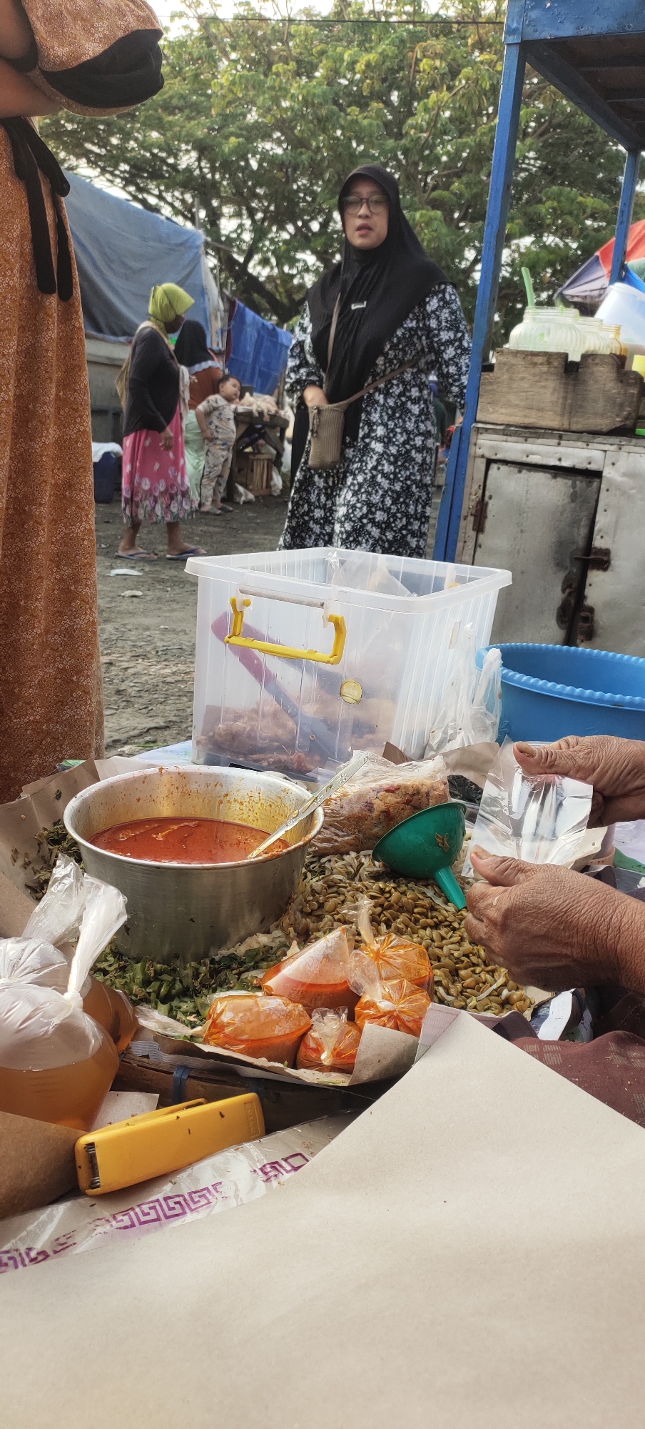 A street food stall in Karangtowo, Indonesia is depicted. A vendor is preparing food, with various ingredients laid out on a table. In the foreground, a pot of red sauce is visible, surrounded by bags of spices and vegetables. A person in the background, wearing glasses and a hijab, appears to be a customer. The stall is set up outdoors, with a blue awning and trees in the background.