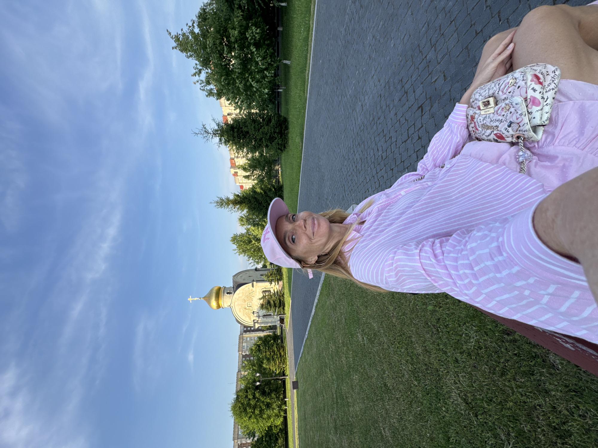 A woman wearing a pink striped shirt and a pink baseball cap sits on a park bench in Kirishi, Russia. A small, ornate church with a golden dome and cross is visible behind her, with residential buildings and trees in the background. The woman is holding a purse adorned with a pattern of lips and text. The ground is paved with cobblestones and a grassy lawn is to her left. The sky is blue with scattered white clouds.