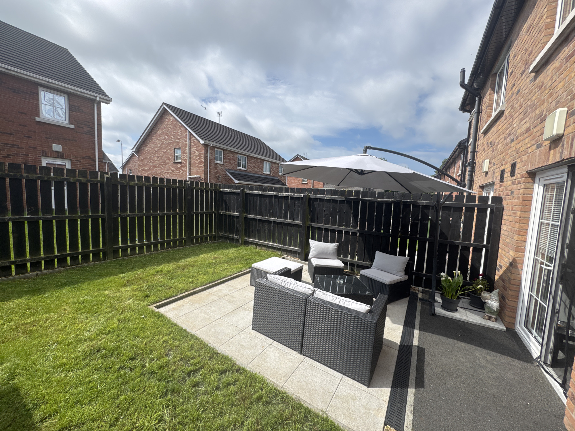 This image depicts a backyard patio area with a lawn and surrounding residential buildings in Craigavon, United Kingdom. The patio features a dark grey rattan furniture set, including a two-seater sofa, two armchairs, and a coffee table with a glass top. Cushions on the furniture are light grey. A large, off-white cantilever umbrella is open and angled over the patio. A black wooden fence encloses the yard. To the left, a well-maintained green lawn stretches towards the fence. In the background, two brick houses with dark tiled roofs are visible, one with white framed windows. To the right of the patio, the brick wall of another house is prominent, featuring white-framed windows and doors, as well as a drainpipe. Several potted plants, including white calla lilies, are placed on a small paved area near the house wall, along with a garden gnome. The sky is cloudy with patches of blue. The overall impression is of a clean and tidy domestic outdoor space.