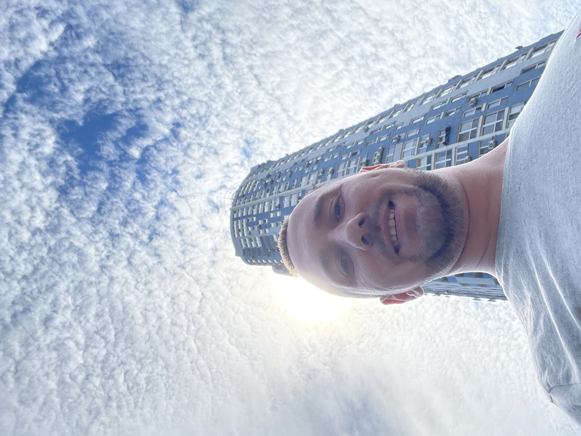 A low-angle, wide-shot selfie is taken from a first-person perspective, featuring a man's head and shoulders against a backdrop of a tall, light blue apartment building and a sky filled with altocumulus clouds. The man, who appears to be in his late 20s to early 30s, has short, light brown hair, a light beard, and is wearing a light gray t-shirt. He is smiling broadly, with his mouth slightly open, revealing his teeth. His head is positioned in the foreground, taking up the lower-left portion of the frame, and his face is tilted upwards towards the sky.
Behind the man, the distinctive light blue apartment building looms large, its facade characterized by rows of white-framed windows and numerous air conditioning units attached to the exterior. The building's architecture suggests a modern, possibly residential structure. The sky is a dominant feature of the image, filled with a dense, textured pattern of white, fluffy clouds that stretch across the entire upper half of the frame. The sun is partially obscured by the clouds, creating a bright halo effect and casting a soft light on the scene.
The perspective emphasizes the height of the building and the expanse of the sky, giving the image a sense of scale. The location is identified as Kyiv, Ukraine, implying an urban setting characteristic of a European city. The image captures a moment of candidness, with the man seemingly enjoying the outdoor atmosphere.