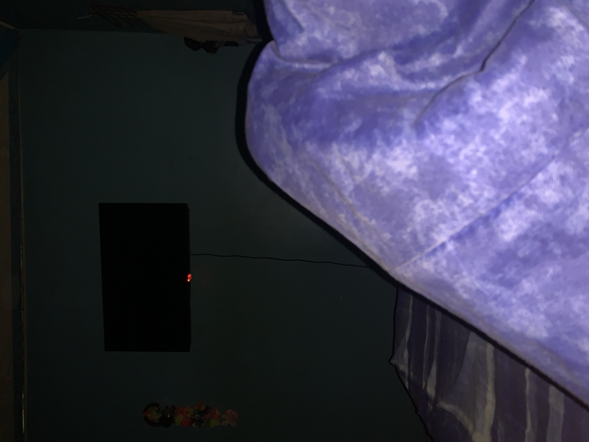 The image depicts a dimly lit indoor room, likely a bedroom, viewed from a slightly high and rotated perspective (appearing as if looking down and to the right from a corner). A significant portion of the frame is occupied by a textured, fluffy purple and white blanket, which appears to be on a bed. Beneath this blanket, another piece of fabric with vertical stripes (purple and white or grey) is partially visible.
The left side of the image features a dark blue or green wall. Mounted on this wall is a rectangular black object, resembling a television or a monitor, which displays a small, red illuminated indicator light on its right edge. A thin black cable extends horizontally from the black object across the wall towards the right. Below the black object, near the bottom left corner of the image, there are several small, colorful decorative items arranged on the wall. The upper left corner of the room shows a ceiling or upper wall area with what looks like draped or hung fabric or clothing items.
The overall lighting suggests it is either nighttime or the room is very dark, with the red light on the electronic device being a noticeable detail. There is no human presence or discernible activity. No text is visible in the image.