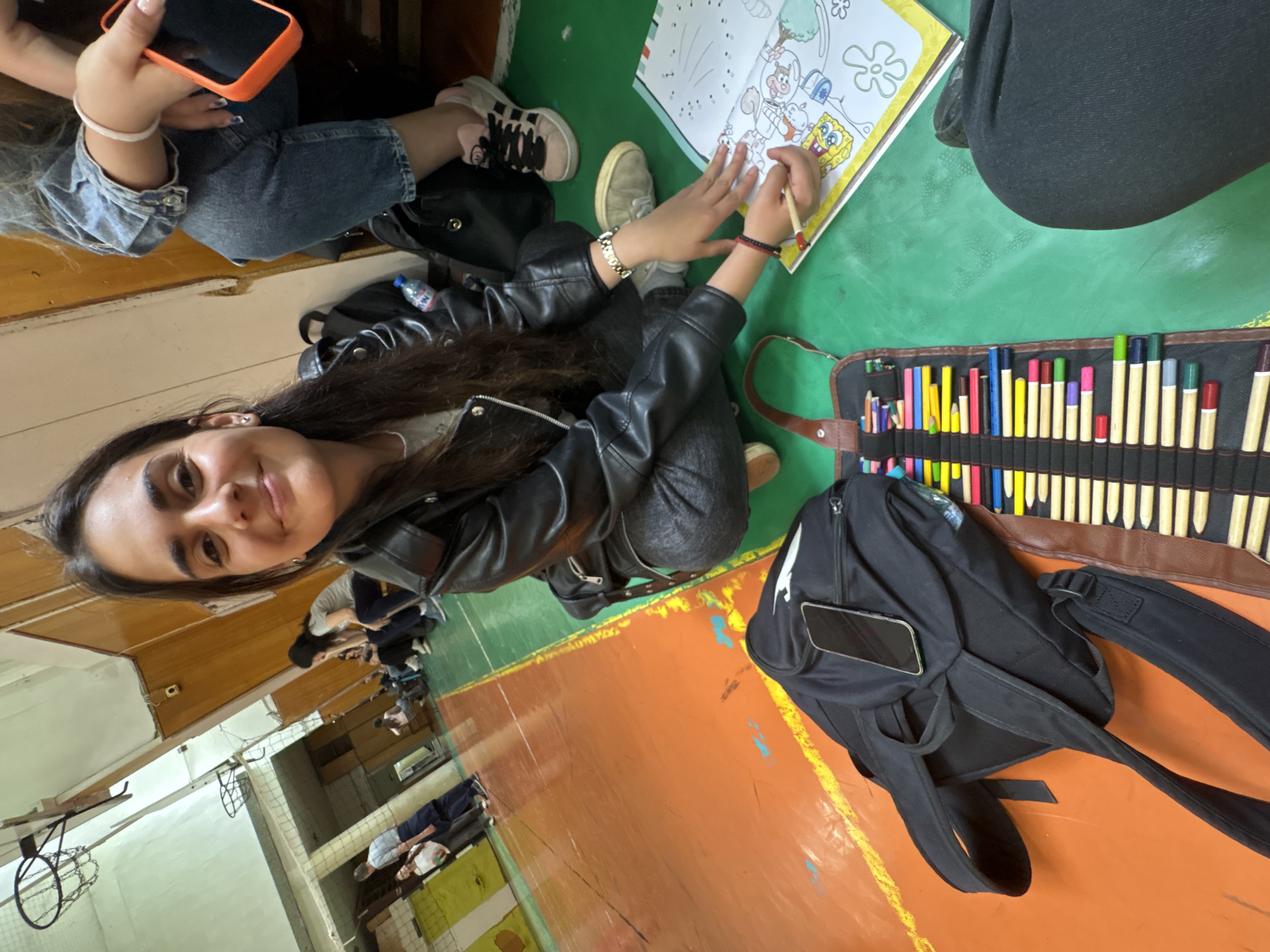 A young woman in a black leather jacket is seated on a green floor, coloring in a "Spongebob Squarepants" coloring book. A pencil case with colored pencils, a black backpack with a phone in its outer pocket, and the lower bodies of other people are present in the scene. The location appears to be a gymnasium, possibly in a school or recreational facility based on the basketball hoop, other seated people visible in the background, and floor markings visible in the background. There is no definitive evidence in the image to confirm the city of Burgas, Bulgaria.