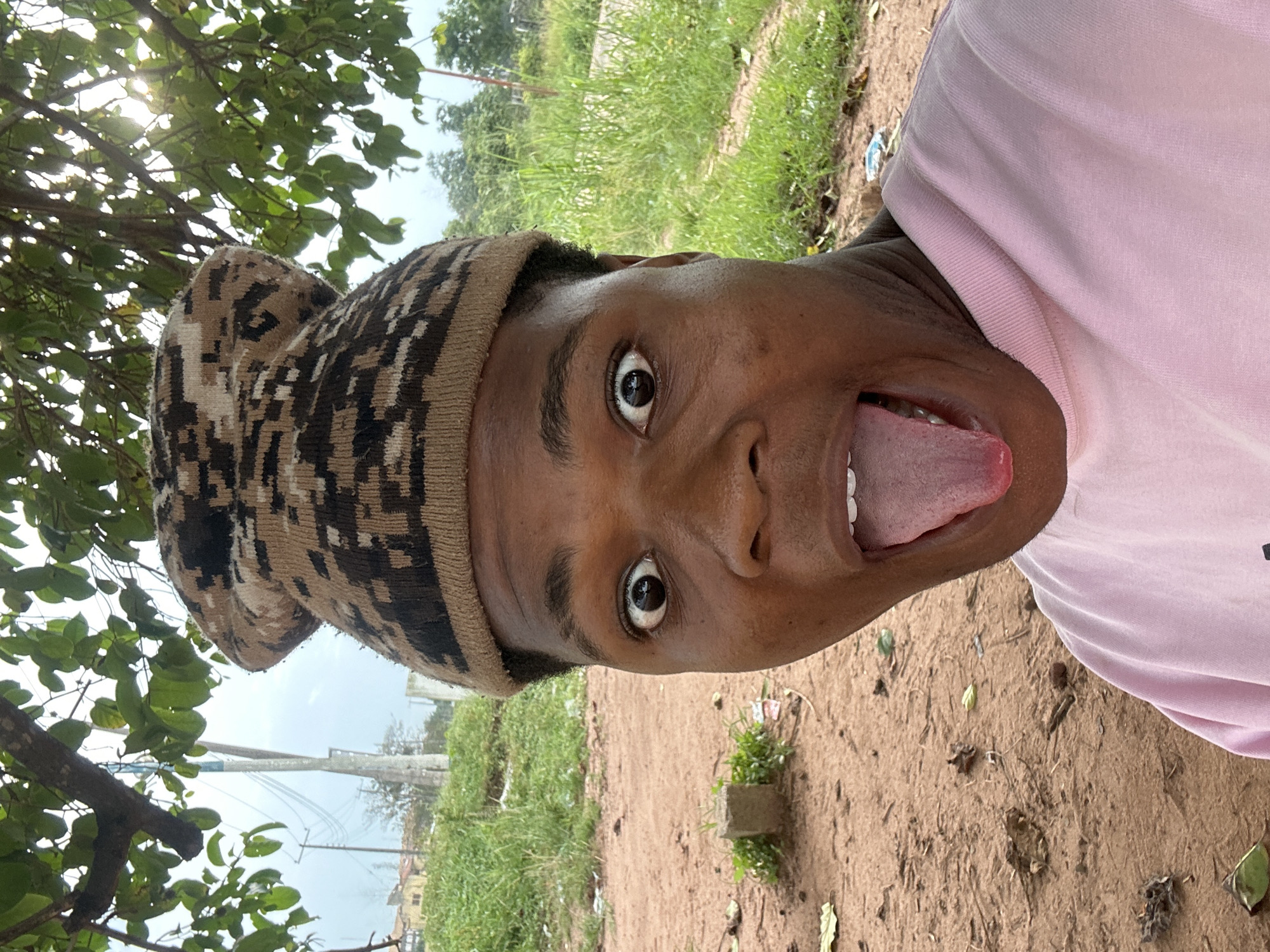 An individual wearing a camouflage beanie and a pink shirt is captured in a selfie outdoors. Their mouth is wide open, with their tongue extended. The background reveals a natural environment with trees, grass, and dirt. In the distance, there are structures resembling buildings and utility poles, suggesting an urban or semi-urban setting. The lighting indicates it is daytime. The geographical context provided is Ihiagwa, Nigeria.