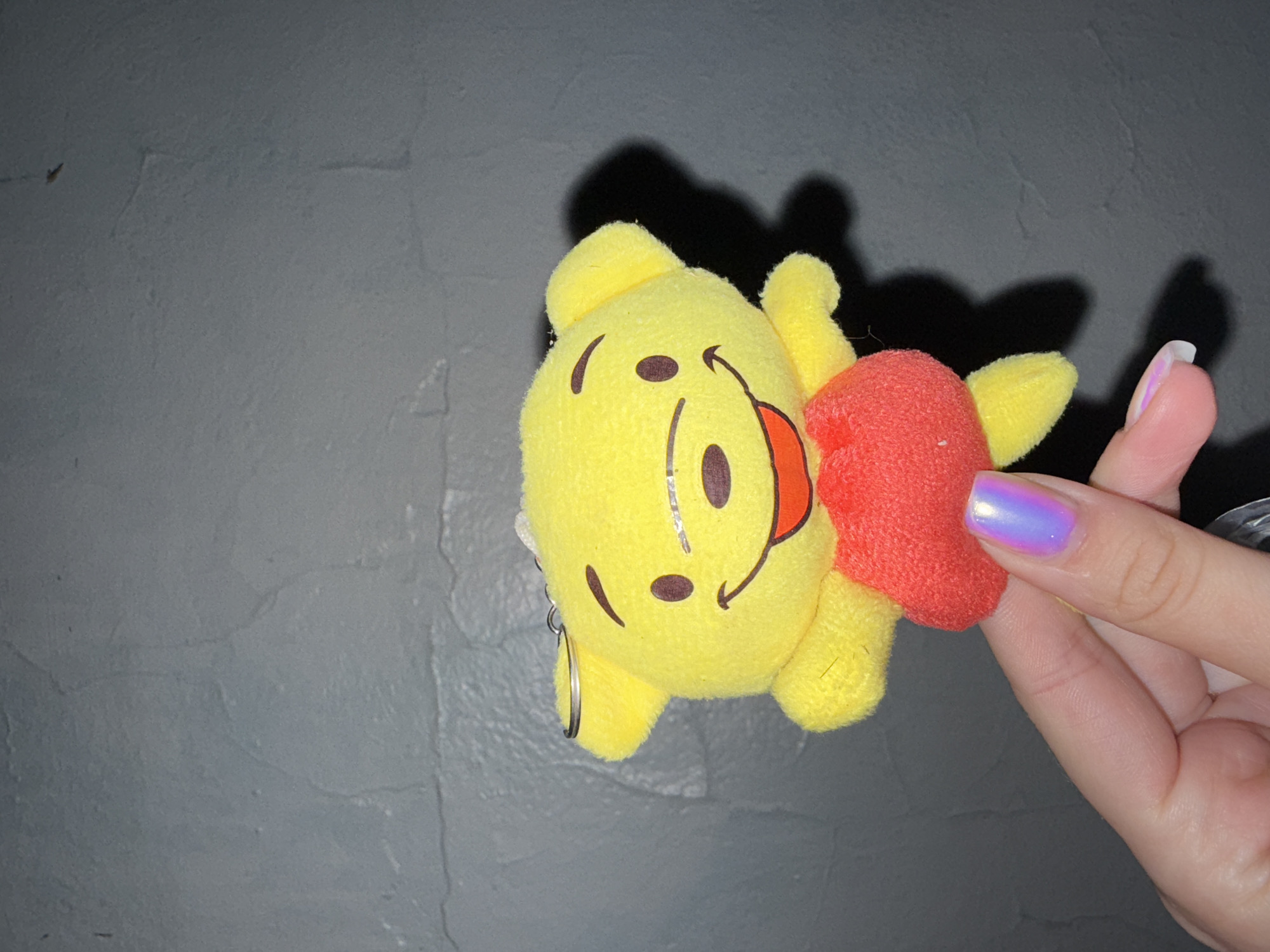 A close-up shot shows a hand with iridescent, shimmering purple nail polish holding a yellow plush toy of Winnie the Pooh. The toy has a red shirt and black embroidered facial features: eyes, nose, and a smiling mouth. A metal keychain ring is attached to the top of the toy's head. The background is a dark gray, textured wall. The image is taken in the city of Sosnovskoye, Russia.