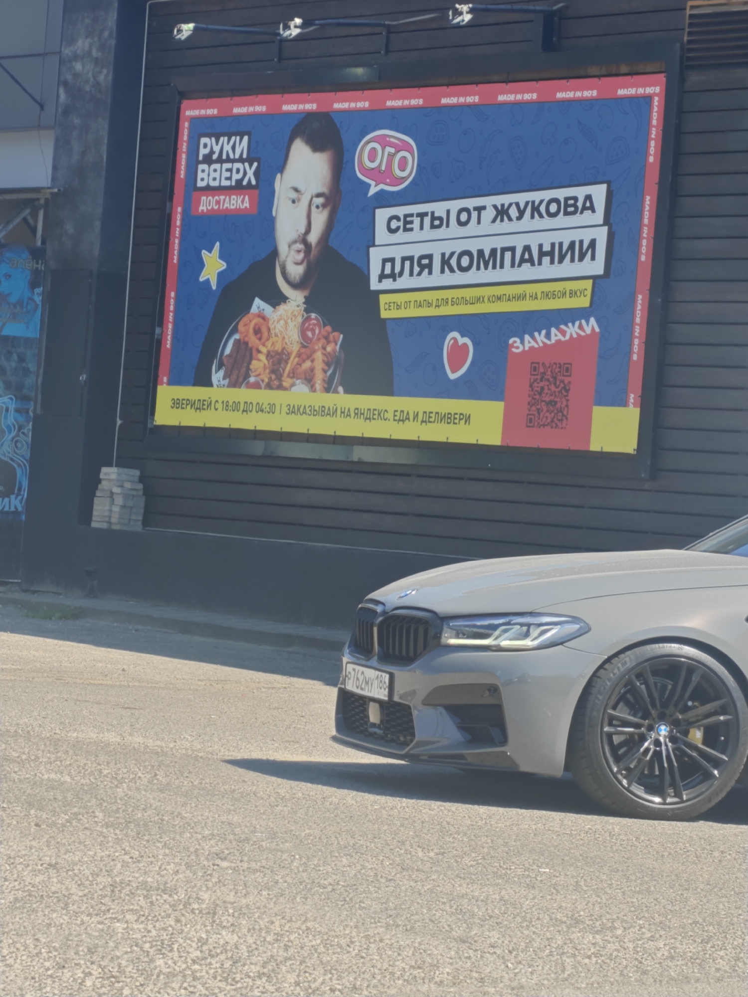 A large billboard is displayed on a dark wooden wall. The billboard features a man with a surprised expression holding a platter of fried food. Text on the billboard reads "РУКИ ВВЕРХ ДОСТАВКА" and "СЕТЫ ОТ ЖУКОВА ДЛЯ КОМПАНИИ" and "СЕТЫ ОТ ПАПЫ ДЛЯ БОЛЬШИХ КОМПАНИЙ НА ЛЮБОЙ ВКУС". There is also a QR code with the word "ЗАКАЖИ" above it. The billboard has "MADE IN 90'S" repeated along the sides. A grey BMW is parked in front of the billboard. The license plate is partially visible, reading "P762MV 186". The scene appears to be outdoors on a sunny day, with a paved road and sidewalk in the foreground. The overall impression is a commercial advertisement for a food delivery service.