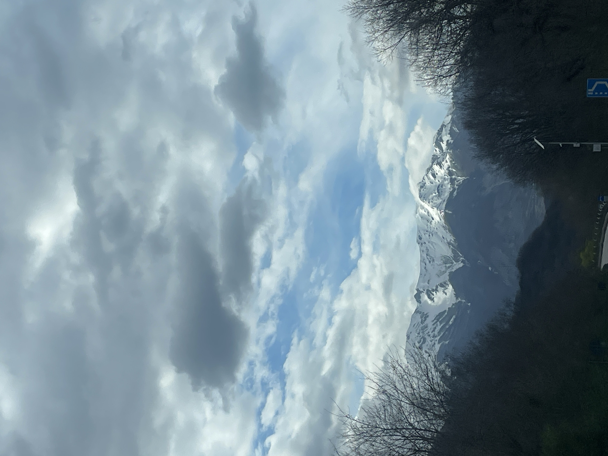 A landscape shot, likely taken from a moving vehicle, shows a snow-capped mountain range partly obscured by clouds. A partly overcast sky with patches of blue is visible. Foreground elements include bare trees and possible road infrastructure, including a blue traffic sign. Location is possibly near San Vincenzo Valle Roveto, Italy.