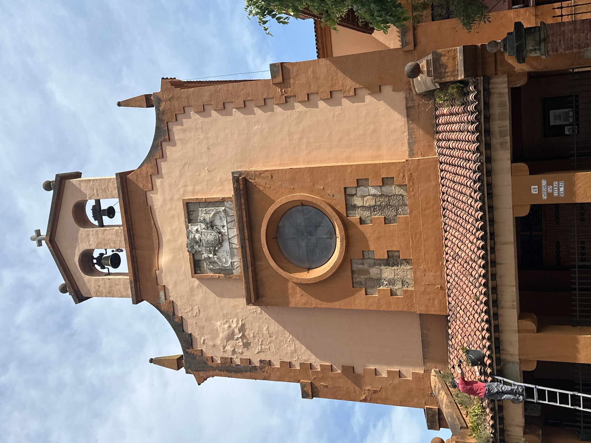 The image shows a building in Gijón/Xixón, Spain. The building is light orange and peach-colored with a terracotta-tiled roof section and a crenellated facade. The architecture appears to be of a historical or religious nature, with a bell tower containing two bells and a cross at its apex. There's a circular window and a carved emblem or coat of arms on the facade.
A man in a red shirt is on a ladder performing maintenance or gardening. He is close to the roof edge. The sky is visible in the background. A sign identifies this as "Parroquia San Esteban del Mar" and the street number as "48."