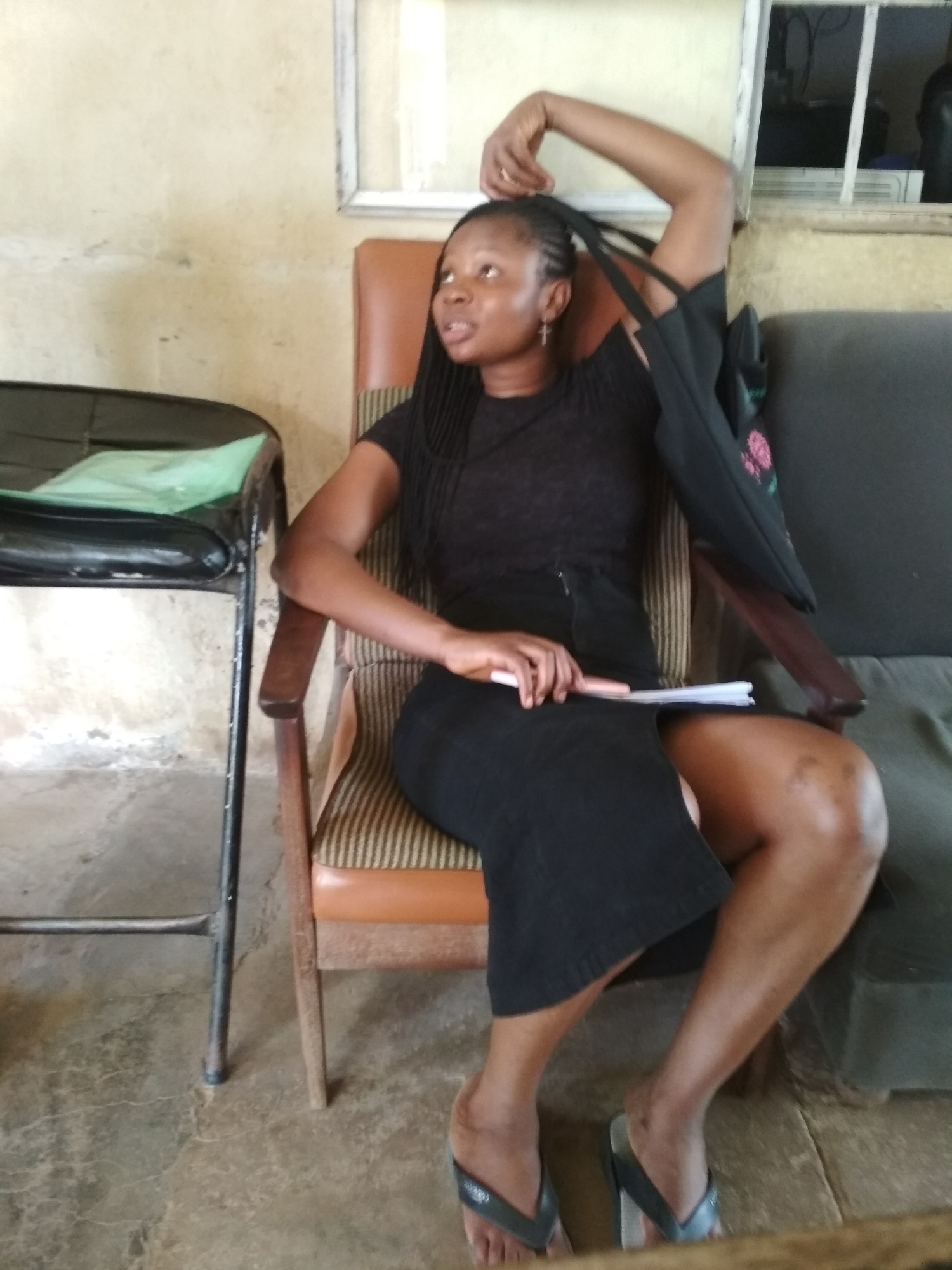 A young Black woman with braided hair sits on a chair in a room in Bisichi, Nigeria. She is wearing a black top and a black skirt, and black flip-flops. She is holding a pen and a notebook in her lap. A black bag is slung over her shoulder. The room appears to be a modest dwelling, with simple furniture and a concrete floor. A window is visible in the background.