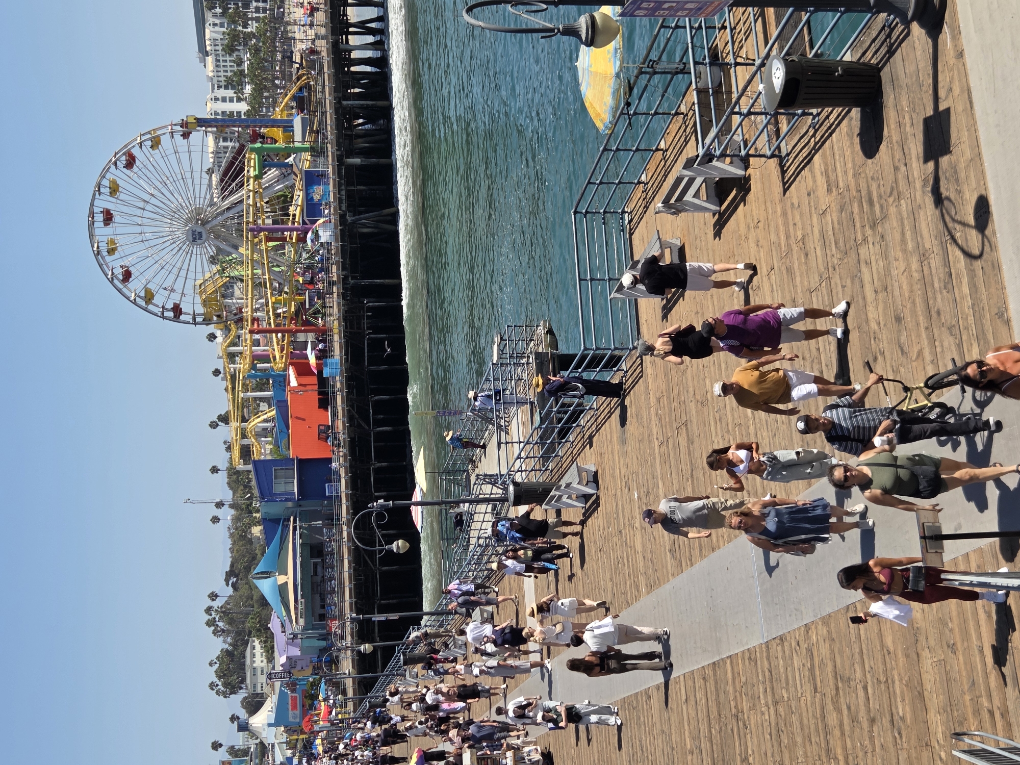 The image shows a bustling scene on the Santa Monica Pier in Santa Monica, California, on a sunny day. A wide wooden boardwalk stretches across the foreground, populated by numerous pedestrians. In the background, the iconic Pacific Park amusement area is visible, featuring a Ferris wheel and roller coaster. The blue ocean water of the Pacific Ocean laps against the pilings of the pier.
People are seen walking in various directions, some individually, others in small groups. They are dressed in casual summer attire, including t-shirts, shorts, dresses, and hats. Several individuals are holding drinks, phones, or bags. Some are engaged in conversation, while others appear to be strolling and taking in the sights. The pier itself is constructed of weathered wooden planks and has metal railings along the edge overlooking the water.
Key objects in the scene include benches, trash receptacles, lampposts, and a yellow and blue striped umbrella providing shade. The environment is characterized by bright sunlight, casting distinct shadows on the boardwalk. The overall atmosphere suggests a popular tourist destination during peak operating hours, with a lively and active ambiance.