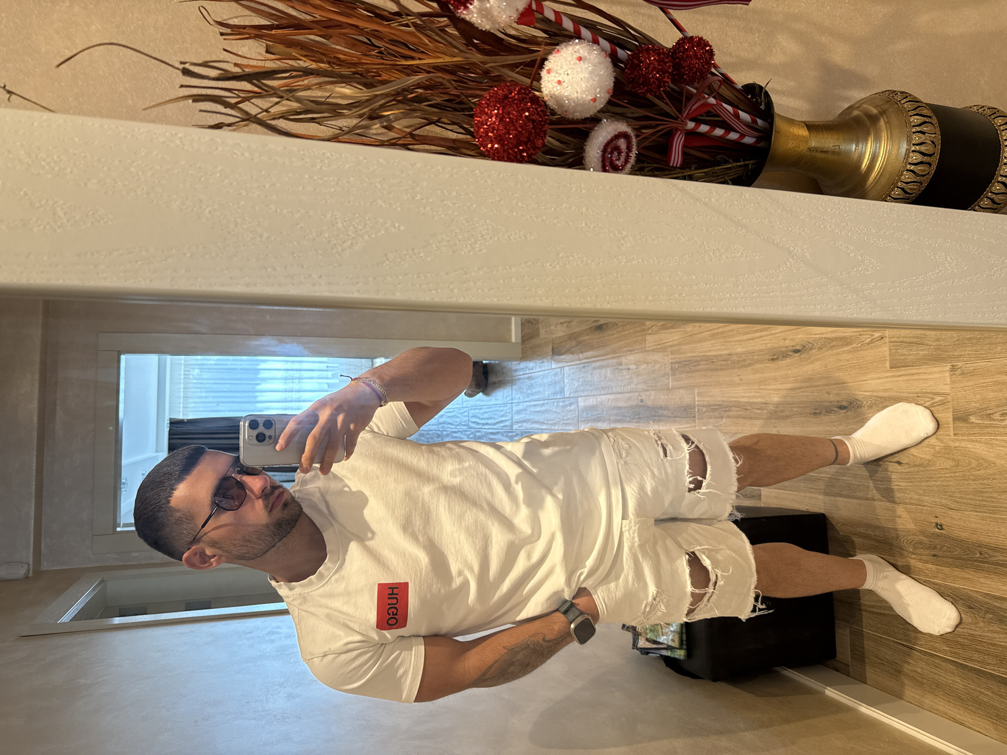 A male stands indoors, facing a mirror, capturing a selfie with his smartphone. He's clad in a white T-shirt with a red "HUGO" logo, and ripped white shorts, completed with white socks. The surrounding environment features wood-patterned flooring and decorative elements, notably a vase containing ornamental branches and red/white baubles placed on a ledge. A suitcase is on the ground next to the man. The image location is presumably within a residence, no definitive evidence is visible to prove a location in the city of Gabrovo, Bulgaria.