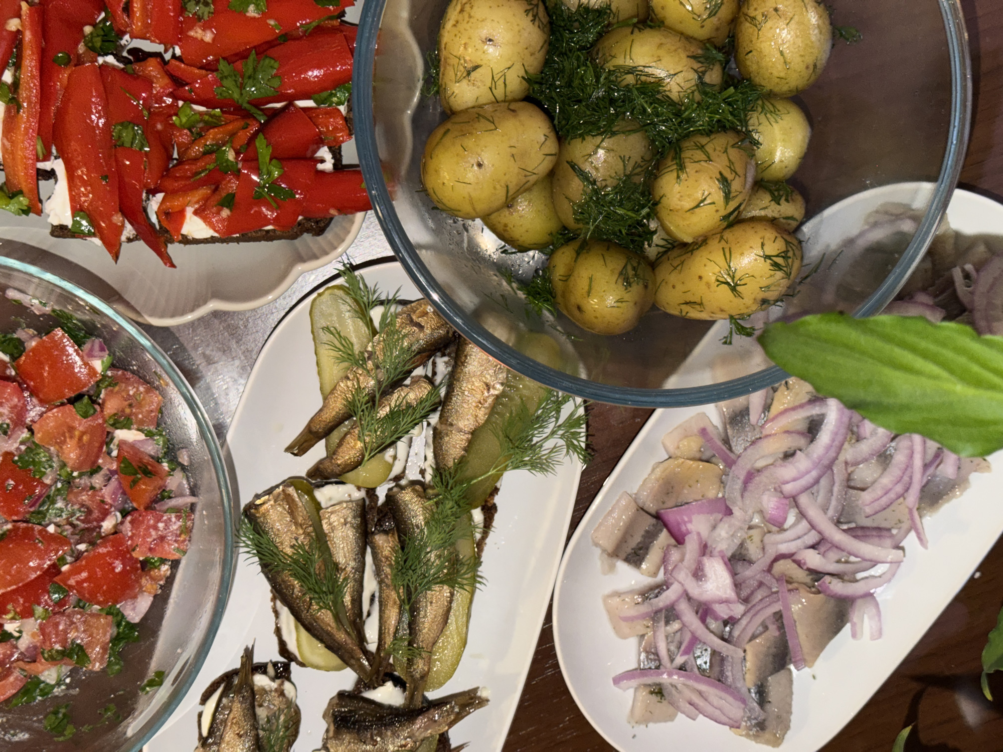 The image shows a table with several plates of food. From left to right, there's a glass bowl with a tomato and onion salad, a plate with brown bread topped with what appears to be red peppers and white cheese, a plate with sprats, pickles, and a white sauce, a glass bowl filled with new potatoes covered in dill, and a plate with sliced herring and red onion. The table is likely in a private home or restaurant setting, but there is no specific information to ascertain that this location is in Budva, Montenegro.