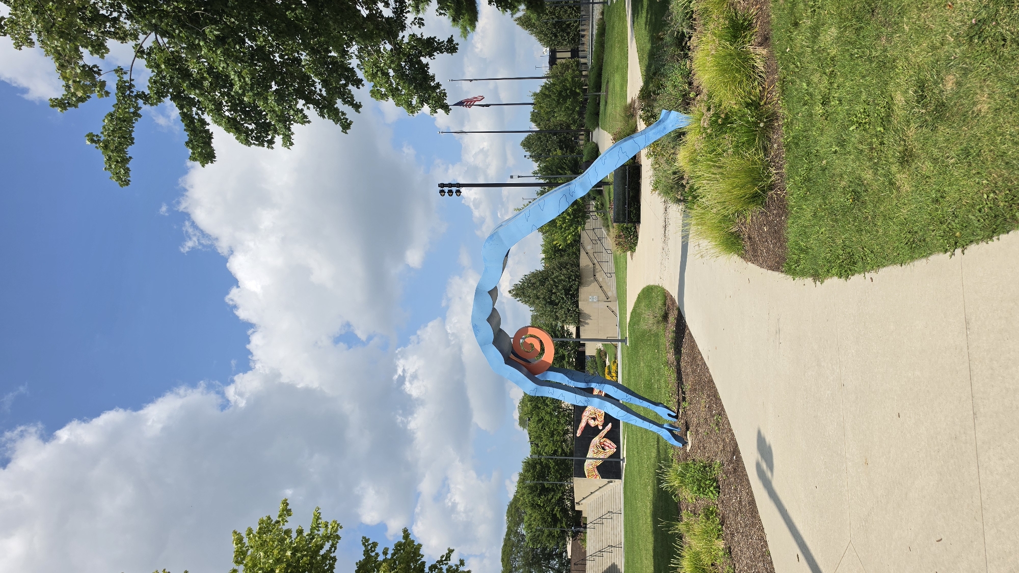 This image captures an outdoor scene with a prominent blue sculpture resembling a person in an arching pose, with an orange circular design at its center. The sculpture is situated in a park-like setting with a paved pathway winding through green grass and landscaped areas with bushes and mulch. In the background, there are tiered concrete stairs leading up to a building, with a mural of hands on a wall visible to the left of the stairs. Several trees with lush green foliage are scattered throughout the landscape, and a few flagpoles with flags are also visible in the distance. The sky is blue with fluffy white clouds, suggesting a sunny day. This sculpture is located at the Wing Park Sculpture Park in Elgin, Illinois, United States of America.