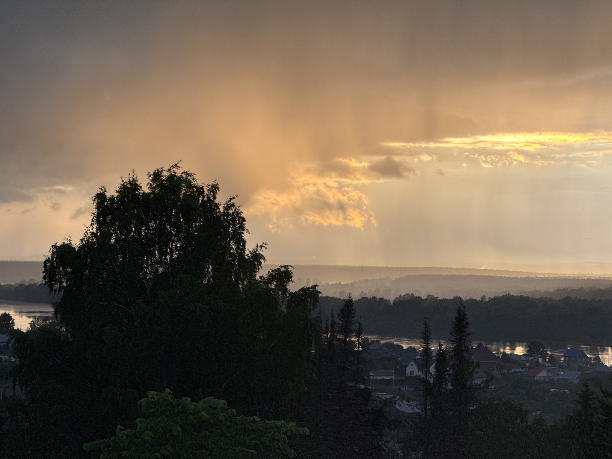 The image captures a dramatic landscape view of the Прикамский region in Russia, likely during either sunrise or sunset, often referred to as the golden hour. The sky dominates the upper portion of the frame, filled with heavy, dark clouds that are spectacularly broken by shafts of golden sunlight. These crepuscular rays pierce through the atmosphere, illuminating parts of the scene and creating a strong contrast, suggesting recent rain or active distant showers. A prominent horizontal band of intense golden light stretches across the mid-sky, where the sun's glow reflects brightly off the cloud formations.
In the foreground and mid-ground, dense clusters of trees, including large deciduous and coniferous varieties, are rendered in dark silhouette against the brighter sky and distant landscape. Their shadowy forms frame the view effectively. Below the tree line, a wide river flows through the terrain, its surface intermittently catching and reflecting glimmers of the golden light. Along the banks of the river, a small rural village or settlement is visible, composed of several houses with distinct rooflines, some appearing white and others colored, such as blue.
The background unfolds in multiple, hazy layers of distant hills and extensive forested areas, which gradually fade into a soft, atmospheric blur. The overall scene conveys a sense of peaceful grandeur and the powerful beauty of nature, characterized by the dynamic interplay of light and shadow. No visible text is present in the image.