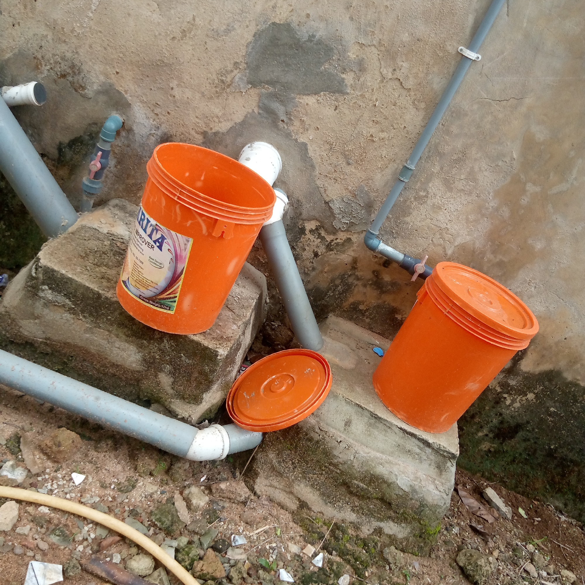 The image depicts an outdoor scene in what appears to be Ogidi, Nigeria, given the style of construction and the product branding on one of the objects. Two orange buckets are visible, one with a lid resting nearby. The bucket on the left has a label that reads "Brita Improver," a dough conditioner. The buckets and lid are positioned on and around concrete blocks. Several gray PVC pipes are running along the wall and ground, some of which are connected with blue fittings and a valve. The background is a textured wall with signs of weathering and repair. The ground is dirt and loose gravel, with some vegetation growing around the concrete blocks. There are no people visible in the image.
