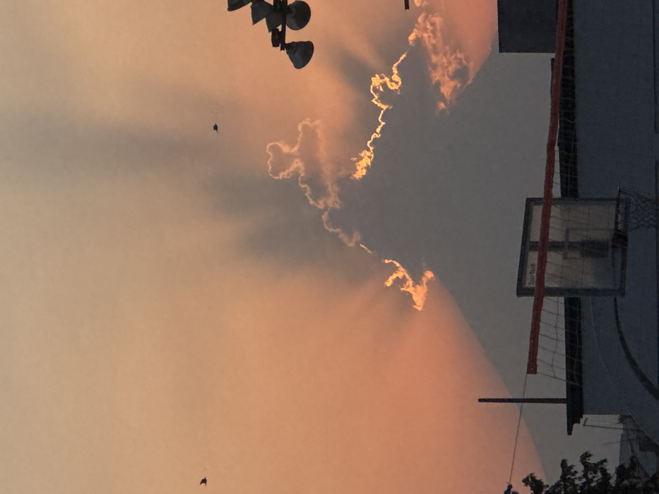 The image displays an evening or morning sky over San Andrés Cholula, Mexico, characterized by a gradient of orange and pink hues. A large, dark cloud mass dominates the center-right, with its edges intensely illuminated by sunlight, creating a golden-orange fringe and prominent crepuscular rays extending upwards and outwards. Two small, dark birds are visible in flight against the sky: one in the upper left quadrant and another smaller one in the upper right. In the foreground, silhouetted against the sky, are portions of urban structures. These include a basketball hoop with a transparent backboard and an orange rim, and an orange-banded volleyball net positioned above it, supported by a vertical pole on the left. A dark structure, possibly a rooftop, underlies these sports elements. In the upper right corner, a portion of a weather instrument, likely an anemometer with cups, is silhouetted.