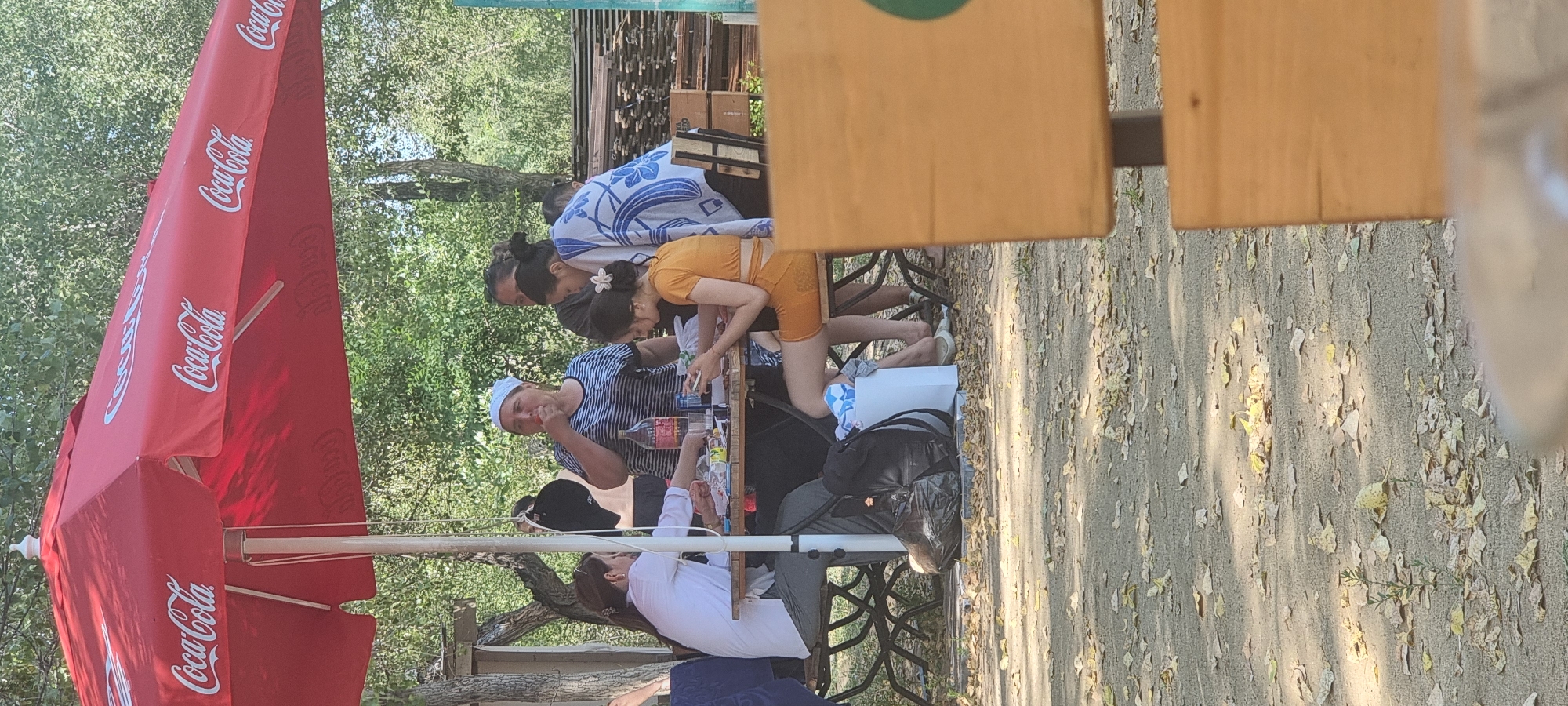 The image shows a group of people sitting around a table under a red Coca-Cola umbrella. There are five individuals visible. One woman in the foreground, wearing an orange top and skirt, is looking at her phone. To her left, another woman in a white shirt and grey pants is also looking at the table. Behind them, a woman wearing a striped shirt and headscarf is eating something. Two other women are standing and looking towards the table. The scene appears to be an outdoor dining area, with trees and foliage in the background. The ground is covered with dirt and fallen leaves. There are also some discarded items like a black trash bag and a white box on the ground. The image quality is moderate, with a slightly grainy appearance. The lighting suggests it is daytime. The context provided regarding the city of Aktobe, Kazakhstan, is not visually verifiable from the image itself.