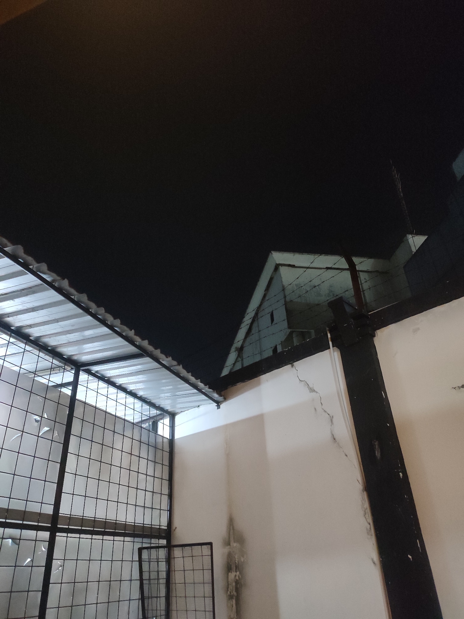 This image depicts a view of a fenced enclosure with a corrugated metal roof, seen from below looking upwards. The enclosure is constructed with black metal framing and wire mesh. Behind the enclosure, a white wall with visible cracks and water stains is present, and further in the background, a building with a triangular roof and barbed wire is visible against a dark night sky. The scene appears to be in an urban environment, possibly a backyard or a secured area. The photograph was taken in Jember, Indonesia.