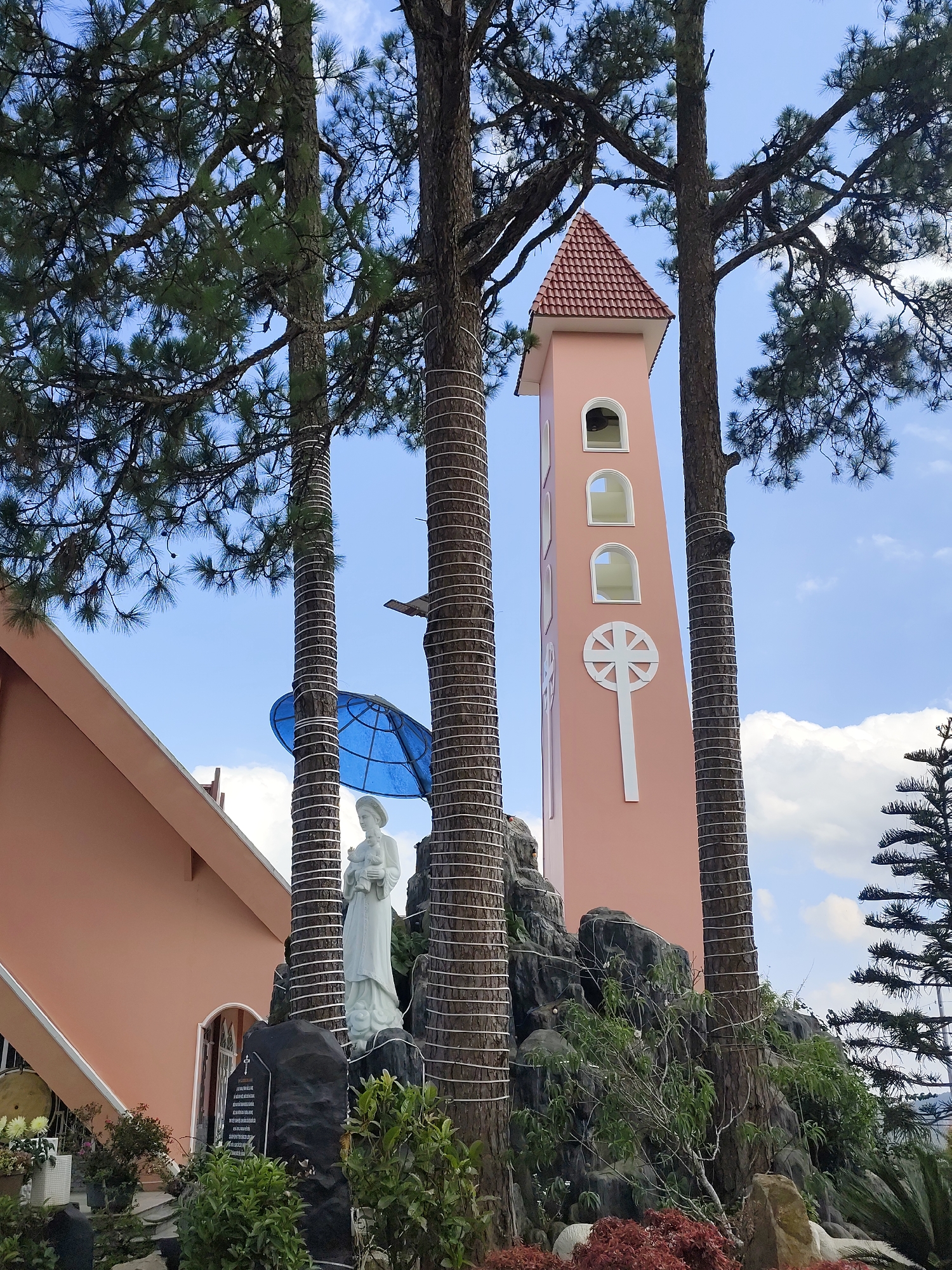 The image captures a view of the Cam Ly Church in the city of Đa Thọ, Vietnam. In the foreground, there is a statue of a woman, possibly Mary holding an infant, set upon a rocky base surrounded by greenery. Adjacent to the statue is a black stone slab with inscriptions. A blue, umbrella-like structure covers part of the statue. The church building is visible to the left with a pink exterior. The most prominent feature is the tall, slender, pink bell tower adorned with a white cross and arched windows. The tower is framed by tall trees that are wrapped with white string. The sky is blue with scattered clouds, and the upper branches of the trees occupy the top portion of the image.
