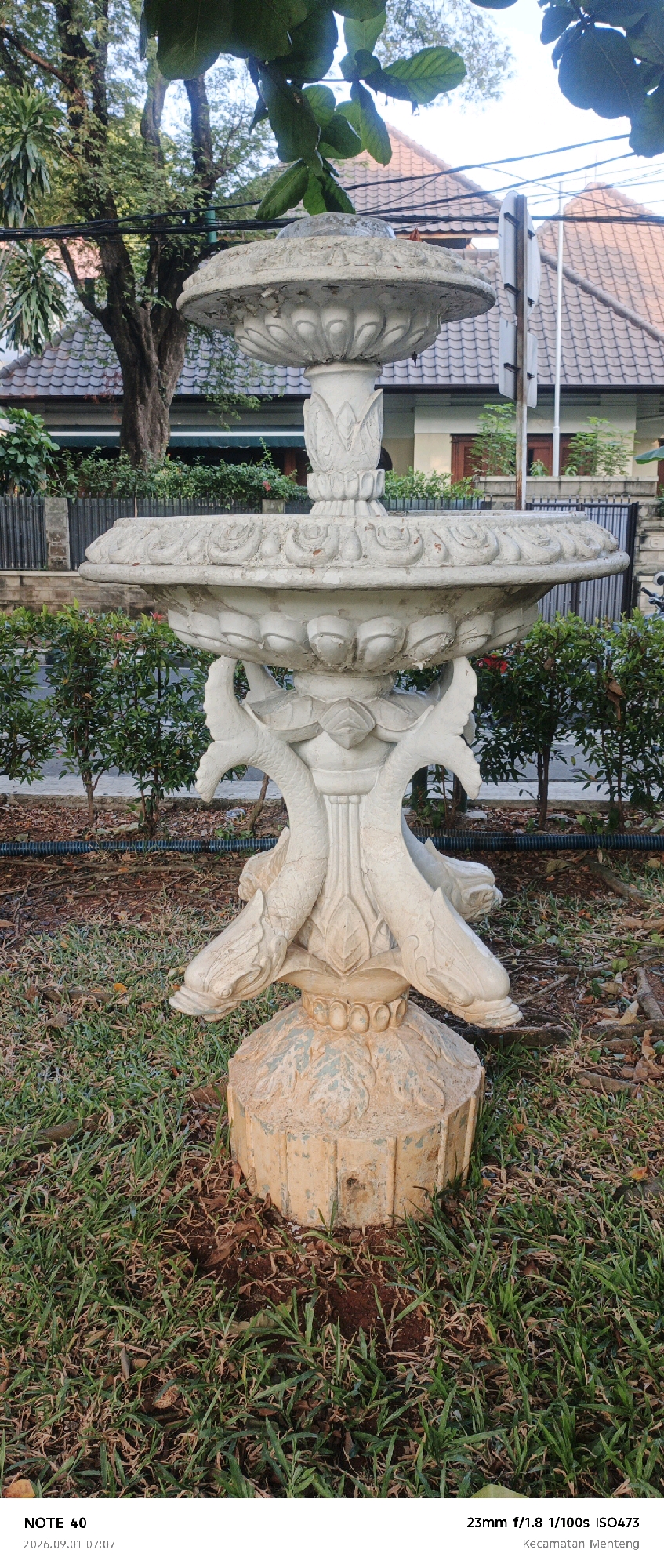 The image shows a multi-tiered stone fountain in a garden setting. The fountain is light-colored, possibly weathered concrete or stone, with decorative carvings of lotus motifs and what appear to be dolphin or sea serpent figures supporting the upper basin. The fountain is centrally placed on a patch of green grass with brown soil and fallen leaves visible around its base. In the background, there are manicured green bushes, a black metal fence, and residential buildings with tiled roofs. Trees with lush green leaves are visible, with some branches extending into the upper part of the frame. The sky is bright, suggesting daylight. The bottom left corner of the image includes text indicating "NOTE 40" and a date and time stamp "2026.09.01 07:07", and the bottom right corner shows camera settings "23mm f/1.8 1/100s ISO473" and the location "Kecamatan Menteng". This suggests the photo was taken in the Menteng district of Central Jakarta, Indonesia.