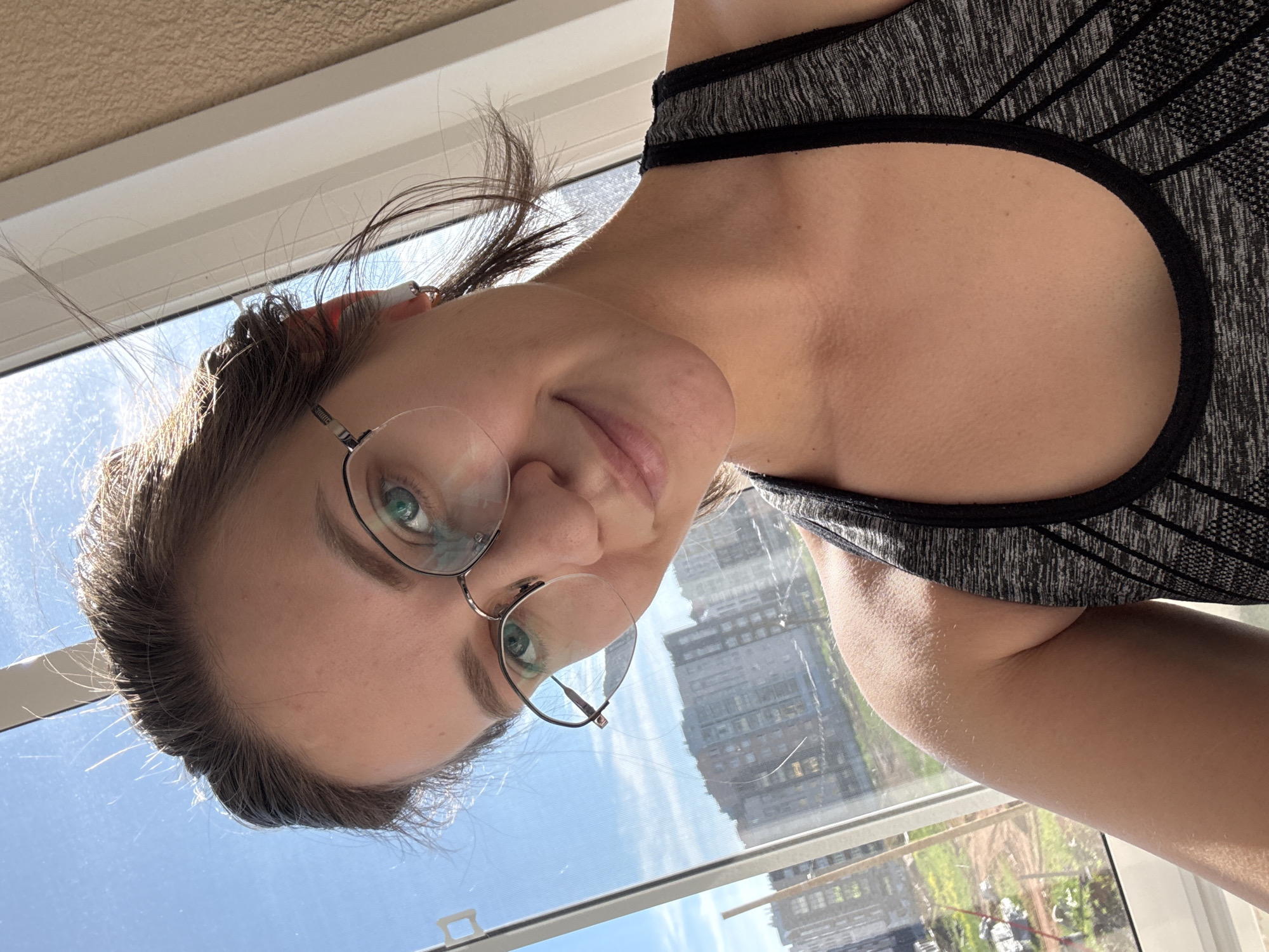 This image is a selfie-style portrait of a young woman, captured from a tilted, slightly upside-down perspective, looking directly at the camera. She has medium-long, dark brown hair, light green eyes, and wears wire-rimmed glasses. A faint, friendly smile is present on her face. A small, white earbud or similar device is visible near her left ear, partially covered by her hair. She is wearing a grey and black patterned sports bra or activewear top.
The setting appears to be indoors, beside a window, through which an urban environment is visible. The sky outside is bright blue with a few scattered clouds, indicating daylight hours, likely morning or afternoon. Through the window, several multi-story residential buildings and some greenery are discernible, characteristic of an urban landscape. The architectural style of the buildings suggests a location such as Pervomaysky, Russia. Natural light streams in from the window, illuminating the woman's face and hair. Reflections of the exterior buildings and sky are visible in the lenses of her glasses. The image conveys a casual and natural moment. No visible text is present in the image.
