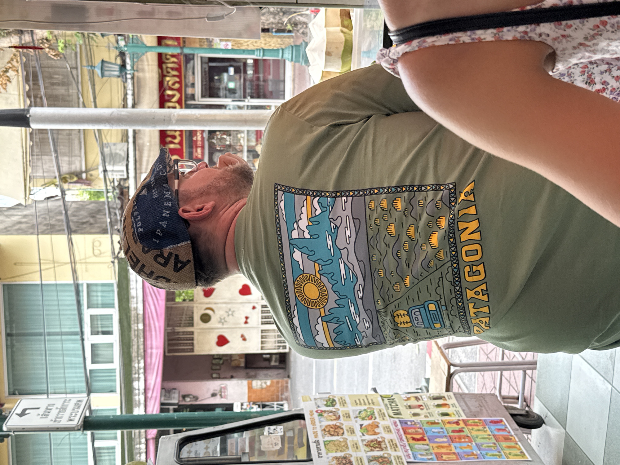 In this image, a man wearing a green t-shirt with a mountain and sun graphic and a patterned flat cap is standing outside an establishment in Bangkok, Thailand. The t-shirt has the brand name "PATAGONIA" printed in yellow letters at the bottom. He is facing away from the camera, looking to his right. To his left, a colorful menu board displays various food and drink options, including "MATCHA" and "SMOOTHIE MENU". Behind him, a street scene unfolds with buildings and overhead wires, typical of Bangkok. A lamppost with a turquoise base and green accents stands to his right, and in the background, two coconuts are visible. A portion of another person's shoulder, wearing a floral-patterned top, is visible in the foreground on the right. The overall scene suggests a casual outdoor setting in the city.