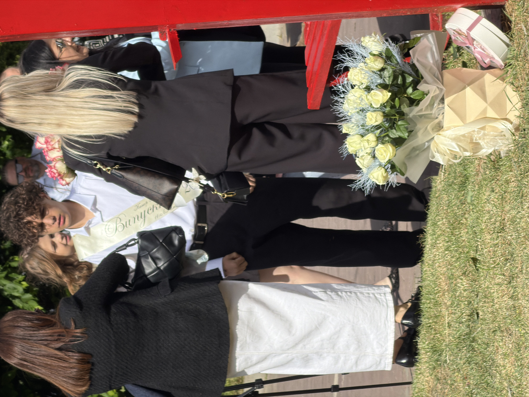 A medium, high-angle shot shows several people standing outdoors. One person has a sash around their chest with the word "Bunyekur" (or a similar word) printed in a Cyrillic script, suggesting an association with graduation. There is a bouquet of light-colored roses next to a geometrically shaped paper box and a small white gift box. The setting appears to be outdoors on a sunny day with grass visible in the background. A red structure, possibly a bench or decorative element, is partially visible at the top of the frame. No elements in the scene confirm the location in the city of Ternopil, Ukraine.