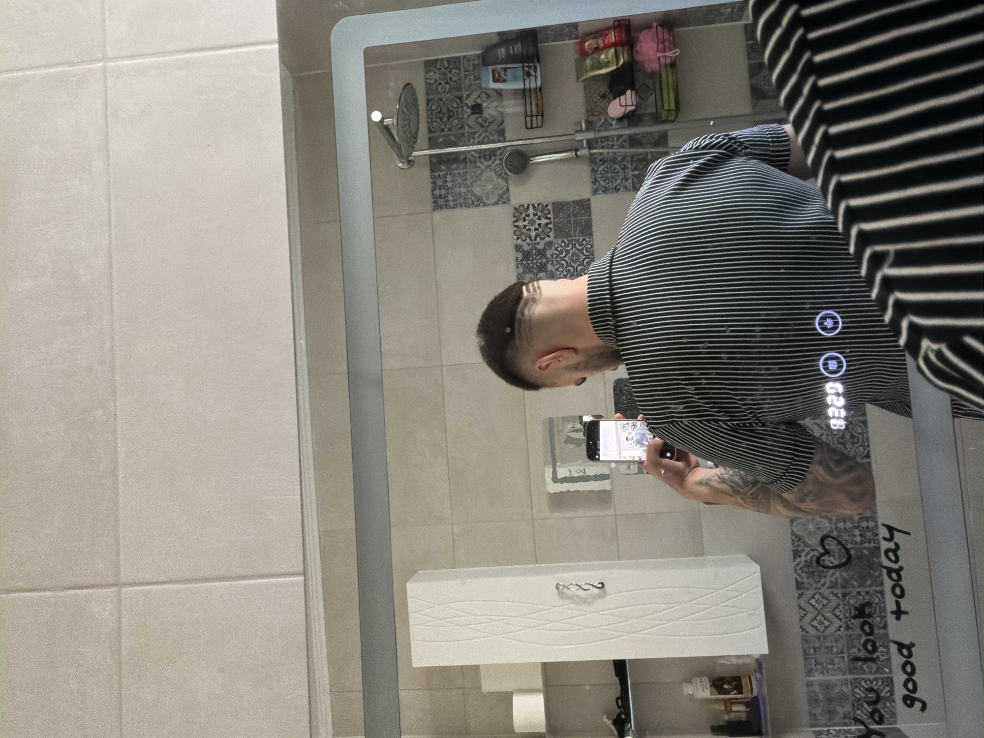 A person is taking a photograph in a mirror. The person has short hair and is wearing a striped shirt. The mirror is in a bathroom with tiled walls. Below the mirror is a white bathroom cabinet. The mirror has text written on it that says "You look good today." The image was taken in the city of Yoakim Gruevo, Bulgaria.