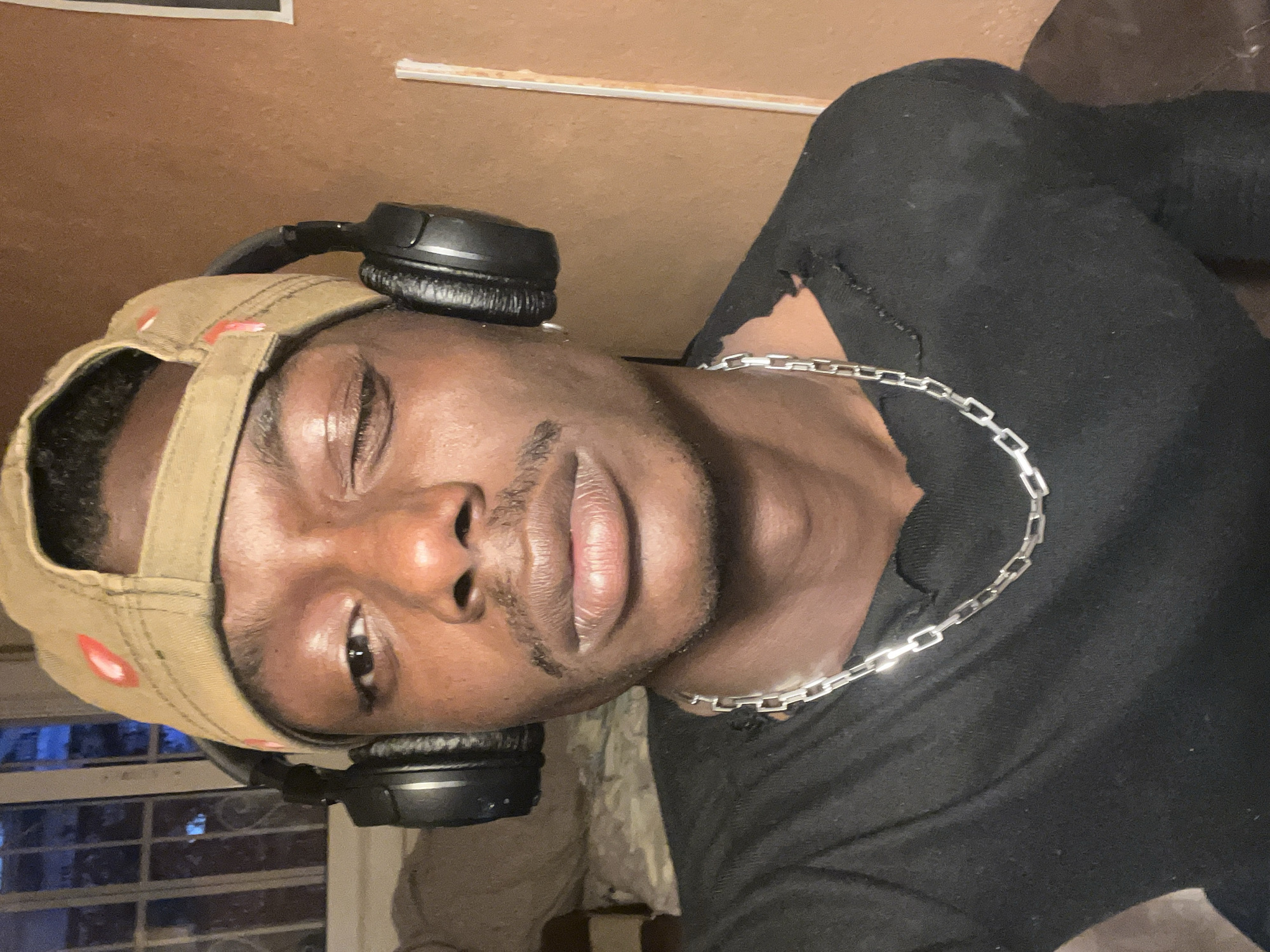 The image, rotated 90 degrees clockwise, features a close-up portrait of a young Black man captured from the chest up. He is looking directly towards the camera with a casual, relaxed expression, his left eye appearing slightly more closed than his right. He has a faint mustache and chin beard.
The man wears a light brown or khaki baseball cap backwards, with subtle red stitching or a design visible on the back strap. Black over-ear headphones are positioned around his neck, resting on his shoulders. He is dressed in a black long-sleeved shirt or sweater, which exhibits visible distressing or tears around the collar and on the upper chest. A silver-colored chain necklace with rectangular links is prominent around his neck, resting on the black fabric.
The background suggests an indoor setting, primarily consisting of a light brown or tan wall/ceiling. In the lower left portion of the rotated image, a window or paneled area with a blueish reflection or view is visible. A thin, light-colored strip, possibly molding or trim, runs horizontally across the upper background. There is no visible text in the image. The lighting appears to be artificial, casting highlights on his skin and the metallic chain.