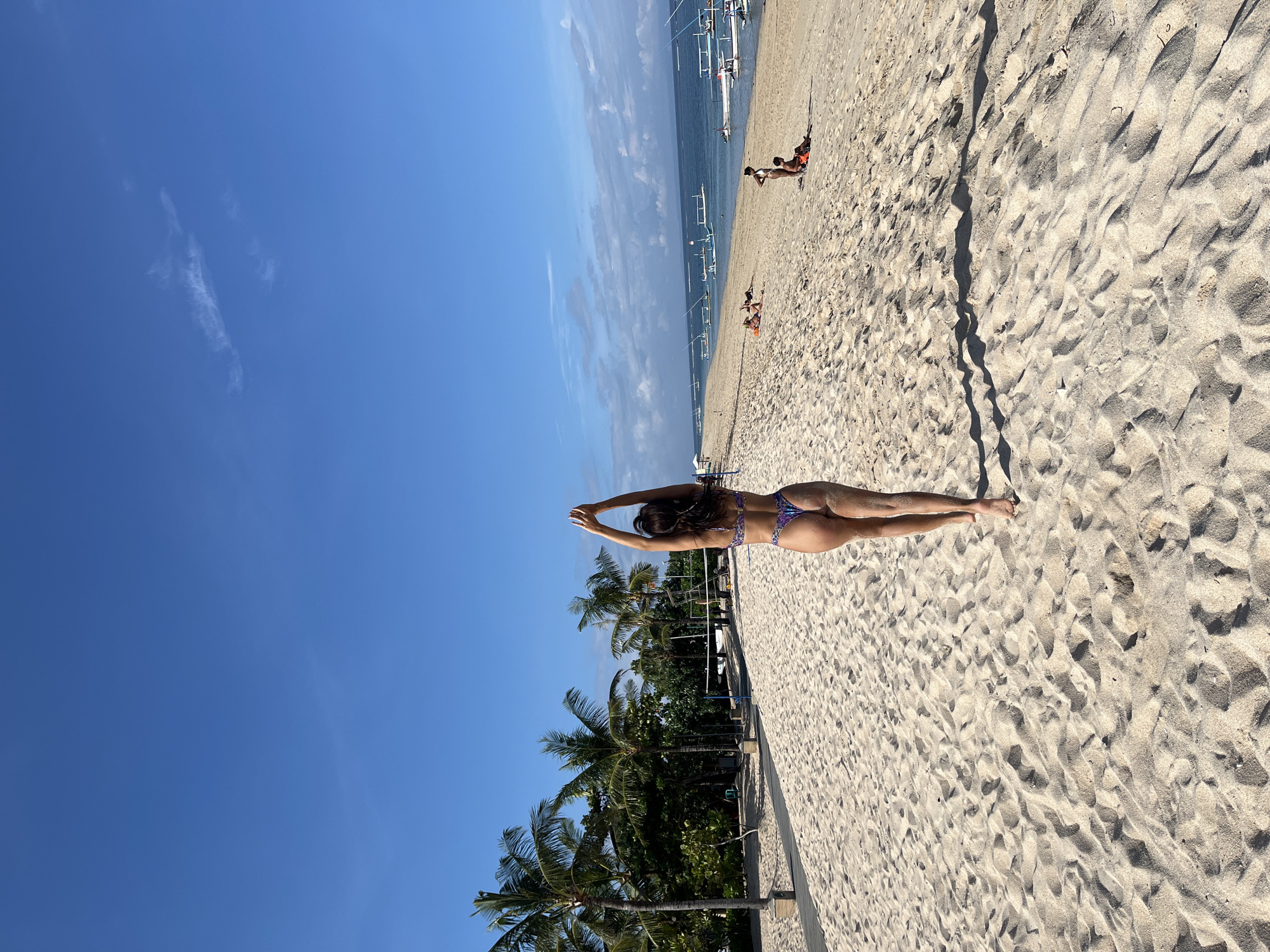 The image captures a vibrant, sunny beach scene, likely in Kuta Selatan, Indonesia, given the context. The primary subject is a woman lying face down on the light-colored sand, stretching her arms above her head. She is wearing a patterned bikini and has dark hair. The perspective is from behind her, looking down the beach towards the ocean.
In the mid-ground and background, the serene blue ocean meets the horizon, with several traditional Indonesian jukung-style boats docked near the shore. Further along the sandy expanse, a few other individuals can be seen relaxing or walking, adding to the leisurely atmosphere. The beach is lined on the left by lush green palm trees, indicating a tropical setting. The sand shows subtle ripples and patterns from the wind and footsteps.
The weather appears perfect, with a clear, expansive blue sky showing only a few wispy clouds. Strong sunlight casts clear shadows on the sand, suggesting it is mid-day. The overall scene conveys a sense of tranquility and a pleasant tropical vacation experience. There is no visible text in the image.
