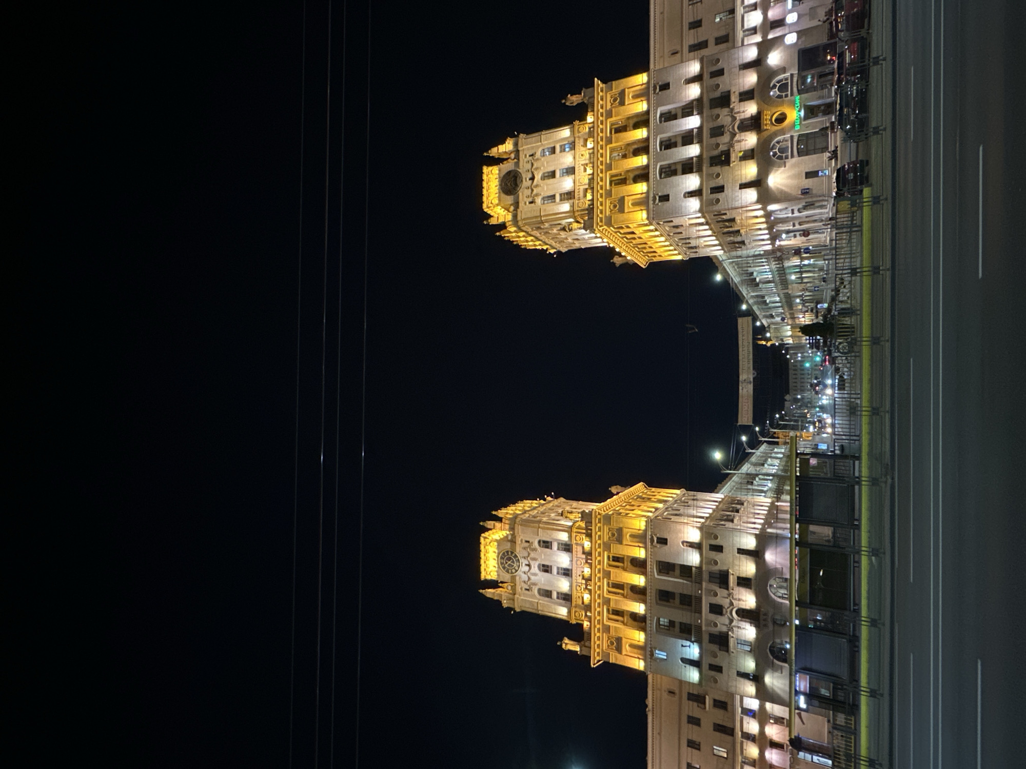 The image shows a nighttime view of the Gates of Minsk in Belarus. The two identical Stalinist Empire-style buildings, illuminated by bright yellow lights, stand symmetrically on either side of a wide avenue. Each building has a prominent clock tower with a round clock face. The foreground features a street with cars and a bus stop. The sky is dark and clear, with power lines visible overhead. The overall scene conveys a sense of grandeur and historical significance of the location.